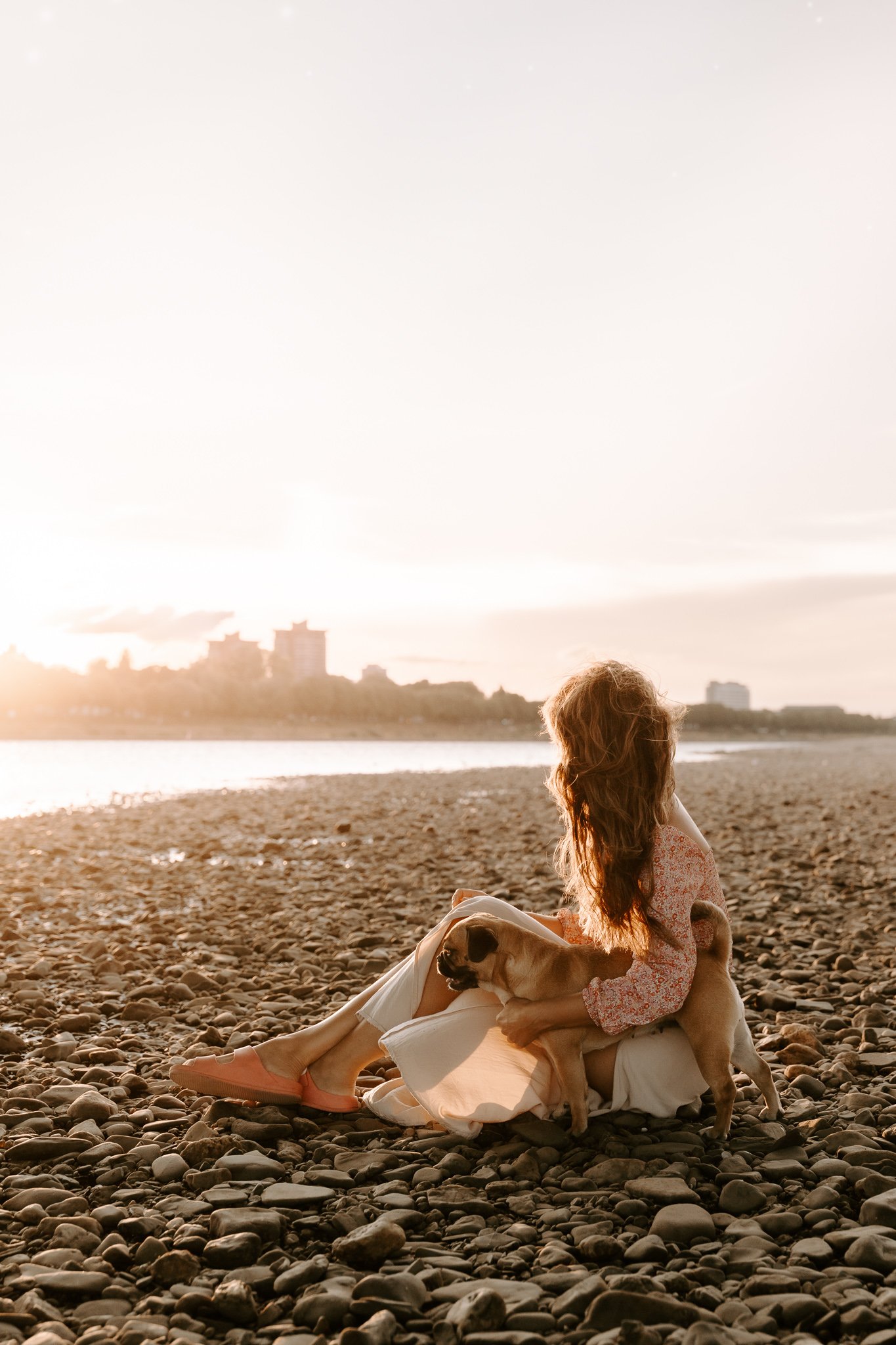 Eine junge Frau sitzt am Rhein in Köln an einem Kieselstrand, umgeben von einem Hund, während die Sonne im Hintergrund untergeht.