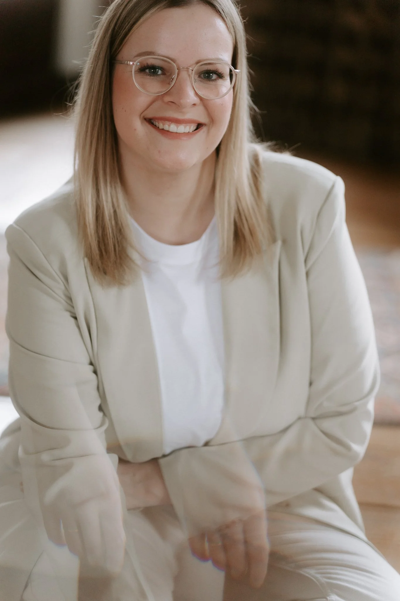 Junge Frau mit Brille, blondes Haar und weißem Outfit, lächelnd, im Innenraum.