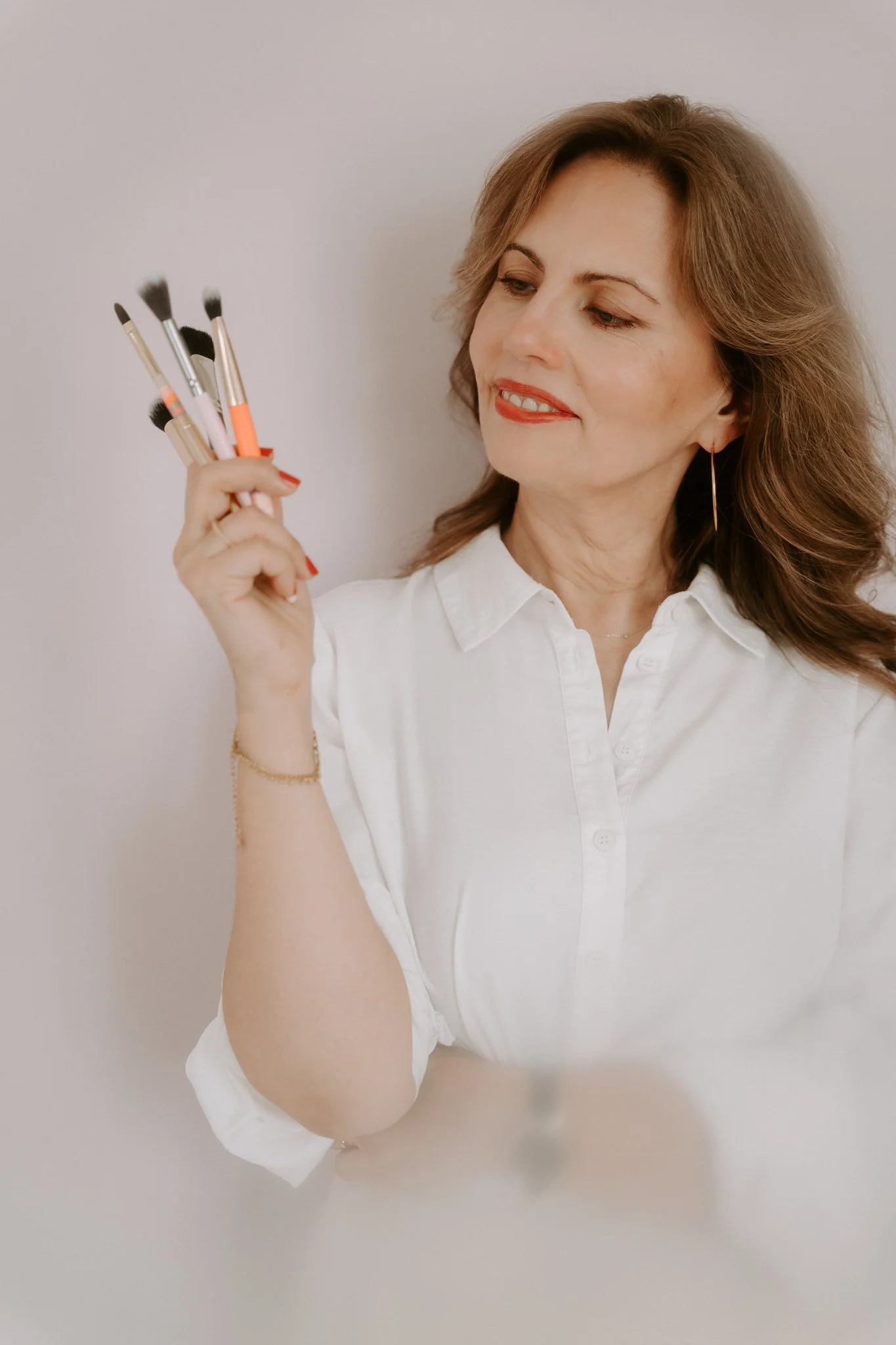 Frau mit roten Haaren und rotem Lippenstift hält mehrere Makeup-Pinsel in der Hand und betrachtet sie.