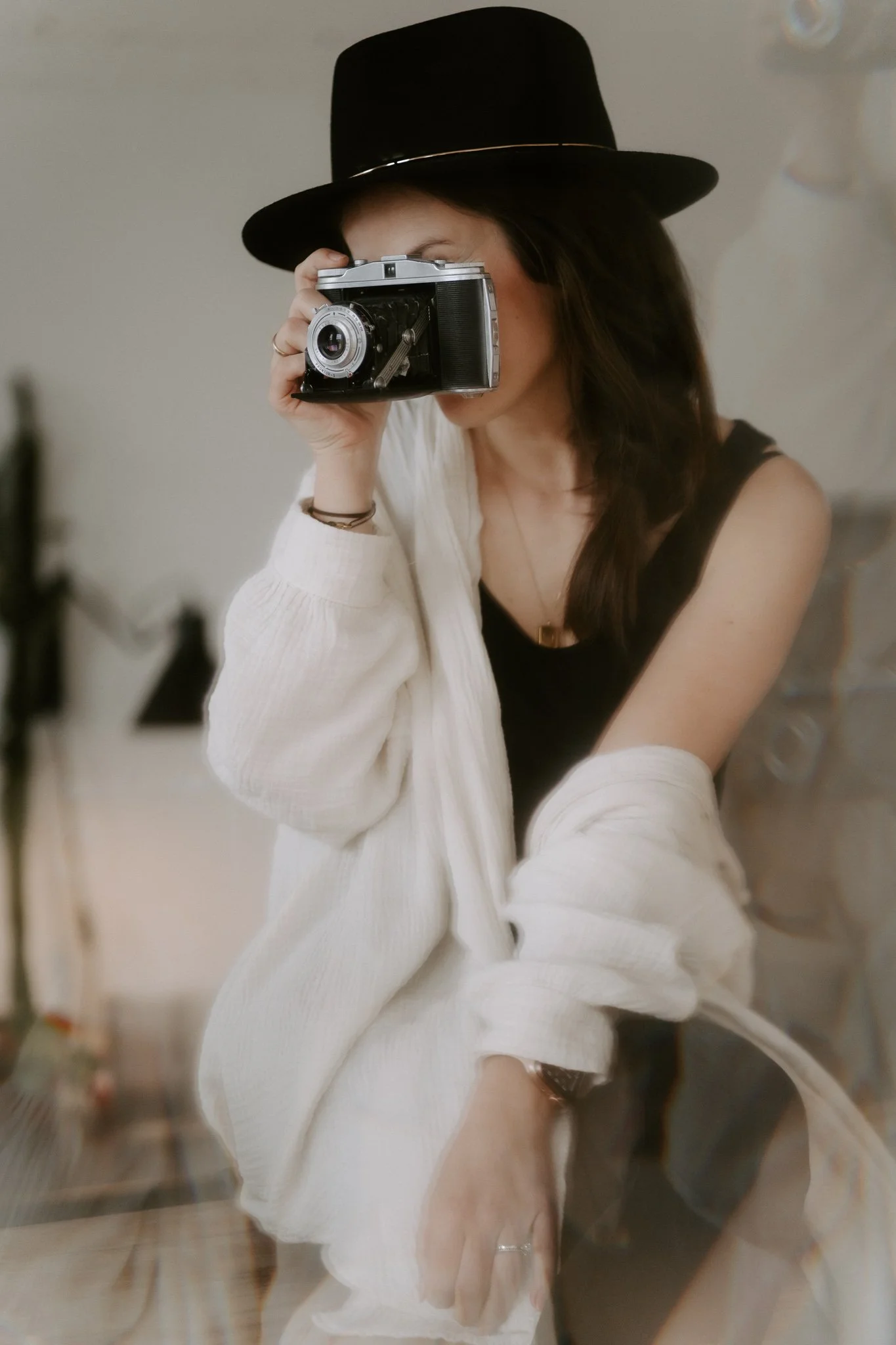 Junge Frau mit schwarzem Hut und Jeansjacke fotografiert sich mit einer alten Kamera, im Hintergrund unscharfe Inneneinrichtung.