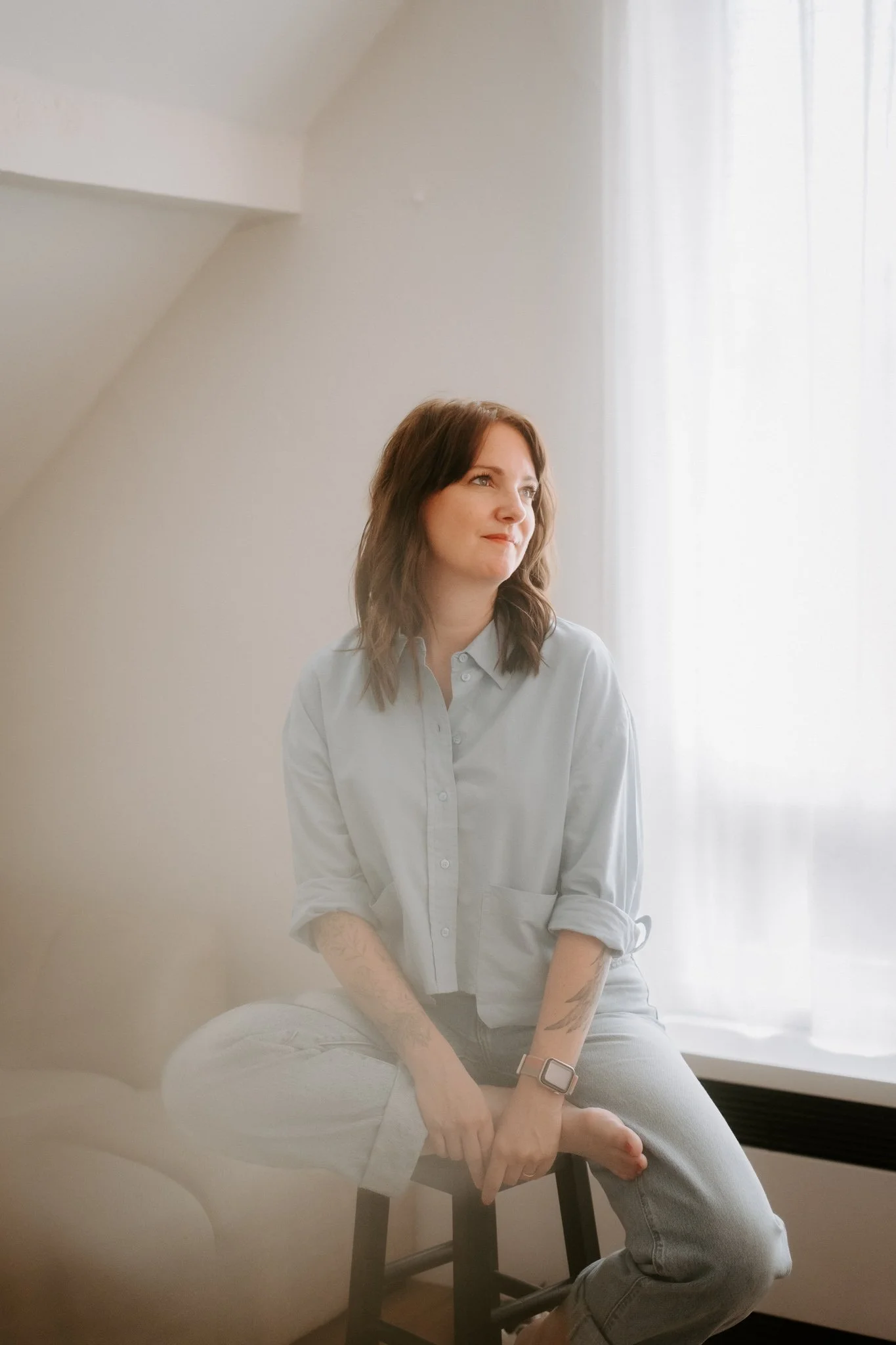 Junge Frau sitzt auf einem Stuhl vor einem Fenster mit weißen Gardinen, trägt ein hellblaues Hemd und Jeans, schaut nach rechts, hat Tattoos am Arm und trägt eine Smartwatch.