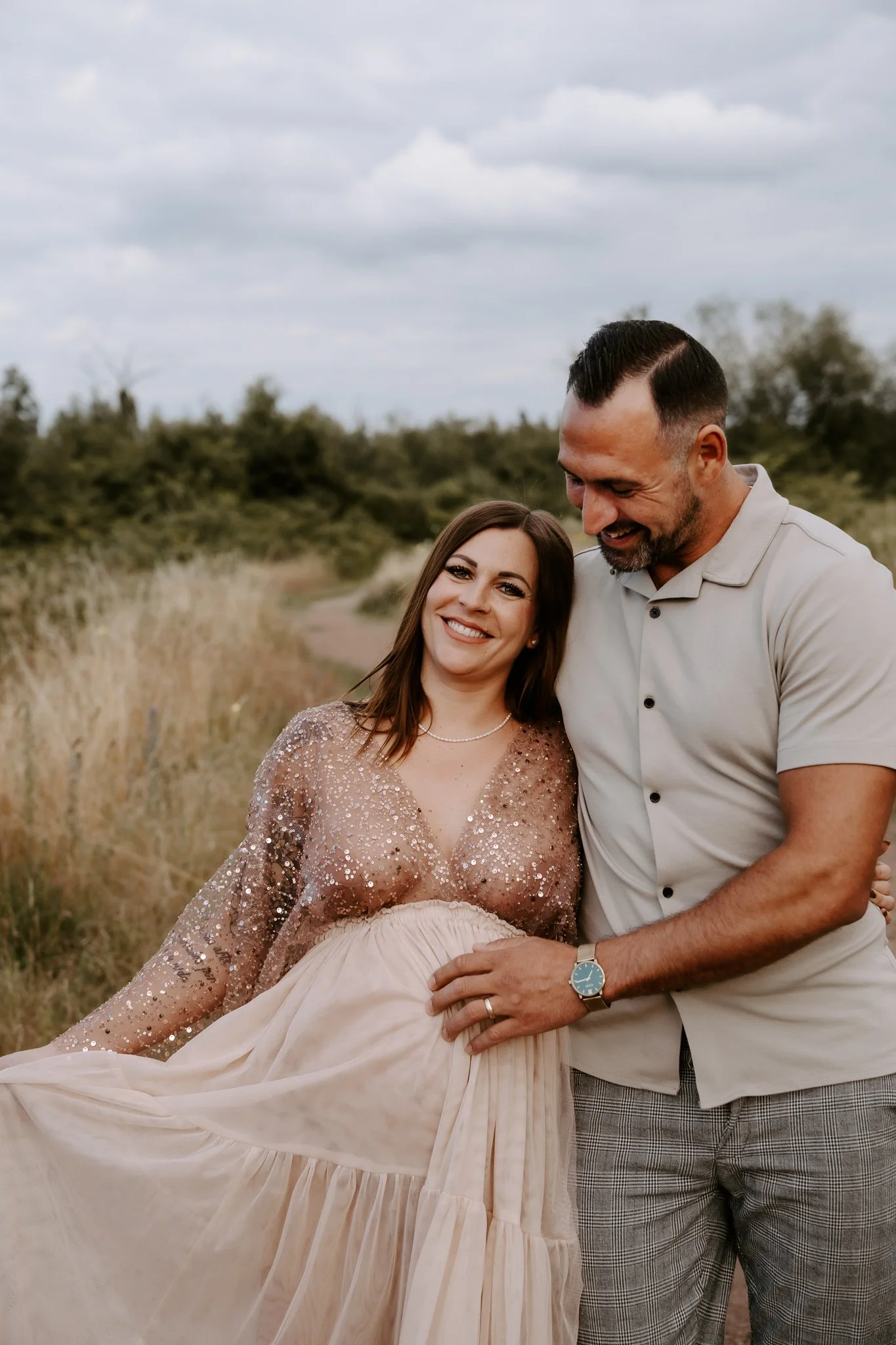 Ein glückliches Paar steht im Freien auf einer Wiese mit hohen Gräsern bei bewölktem Himmel. Die schwangere Frau trägt ein pastellfarbenes, glitzerndes Kleid und lächelt, während der Mann sie liebevoll festhält.