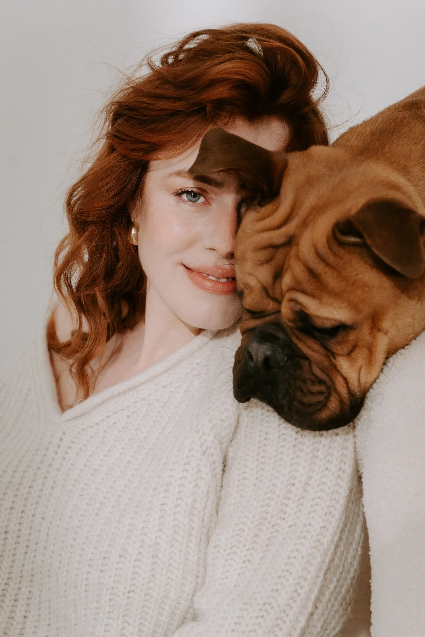 Eine Frau mit roten Haaren und einem weißen Pullover, die mit ihrem Hund kuschelt, der auf einem Sessel liegt