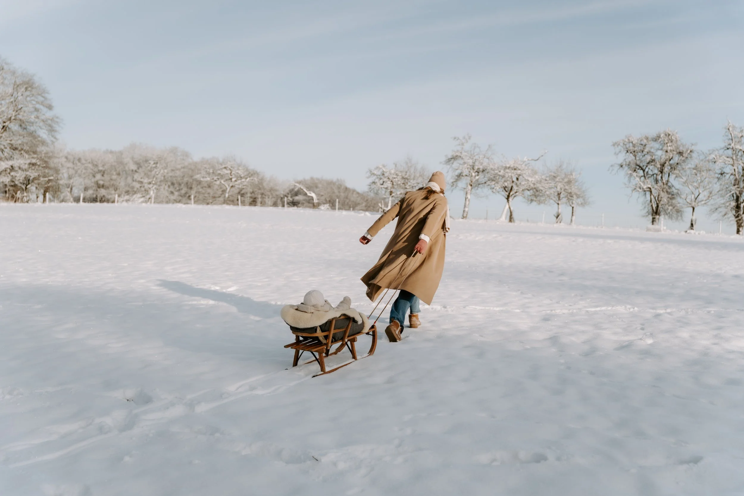 Person zieht ein Kind in einem Schlitten durch verschneites Landschaft mit Bäumen im Hintergrund, sonniger Herbsttag.