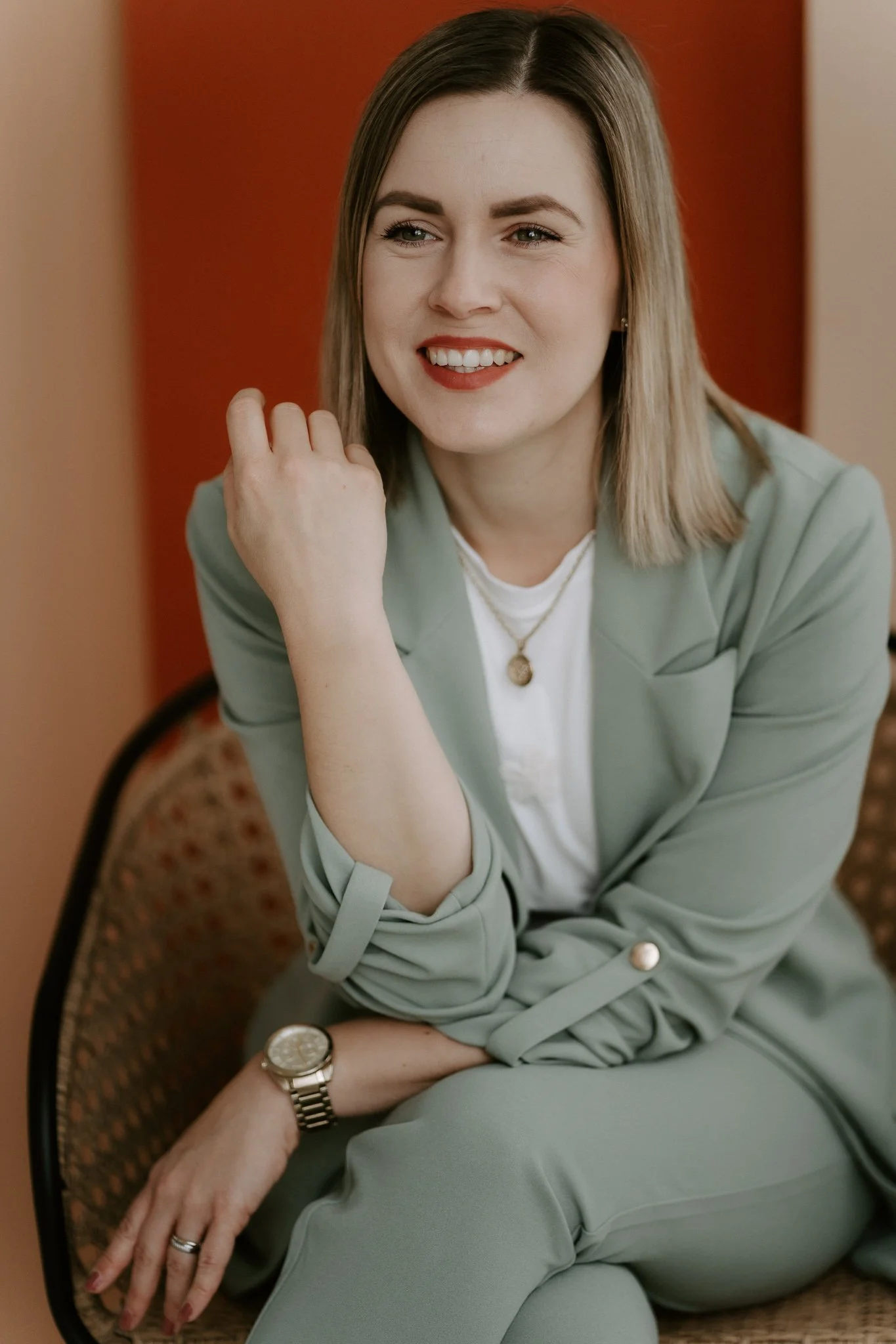 Junge Frau mit blonden Haaren trägt eine grüne Anzug, sitzt auf einer Stuhl, lächelt, trägt eine Uhr und Halskette, vor einem roten Hintergrund.