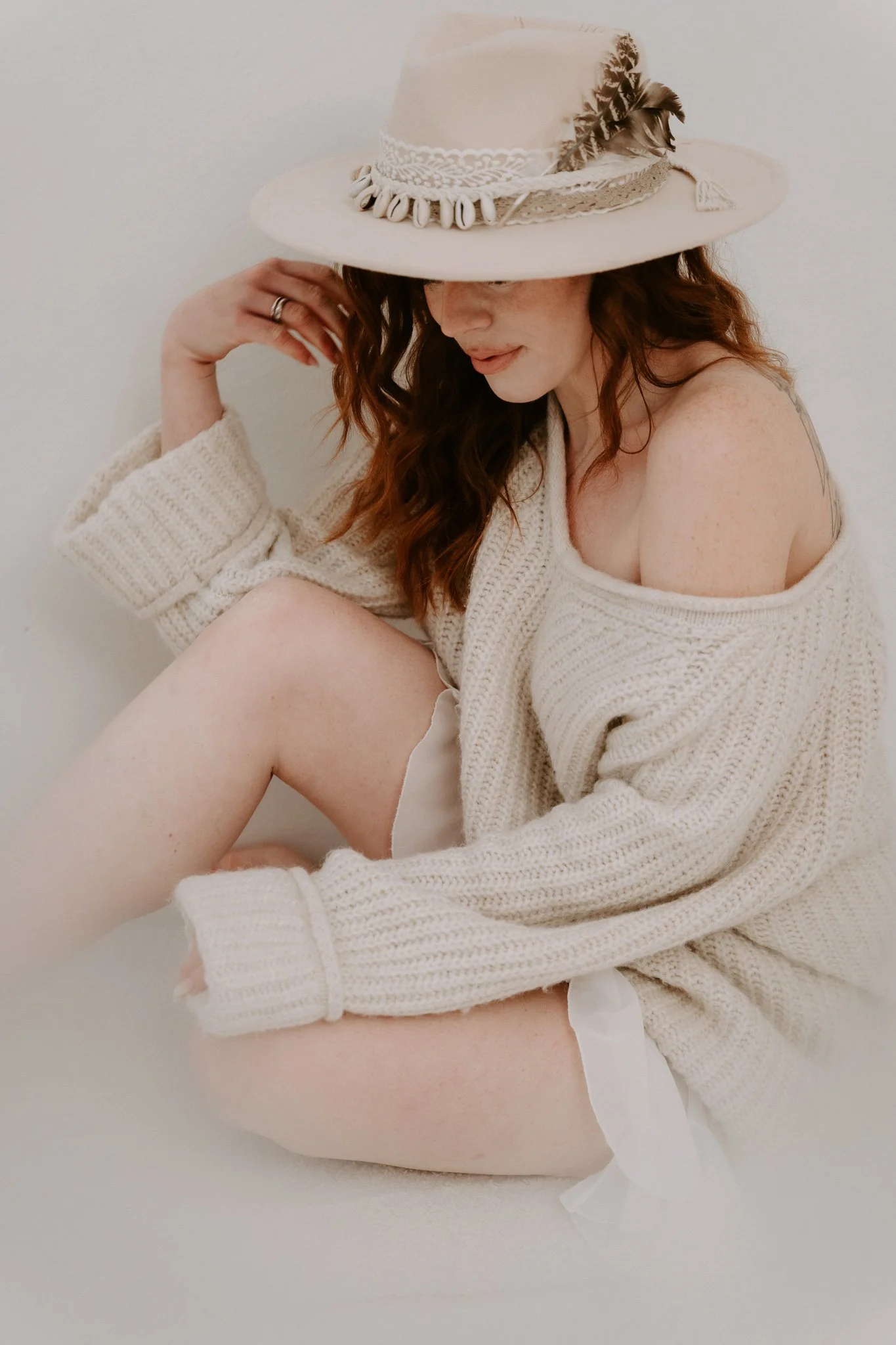 Frau mit roten Haaren, in einem weichen, beige Cardigan, sitzend, trägt einen großen, stilvollen Hut mit dekorativen Elementen, auf neutralem Hintergrund.