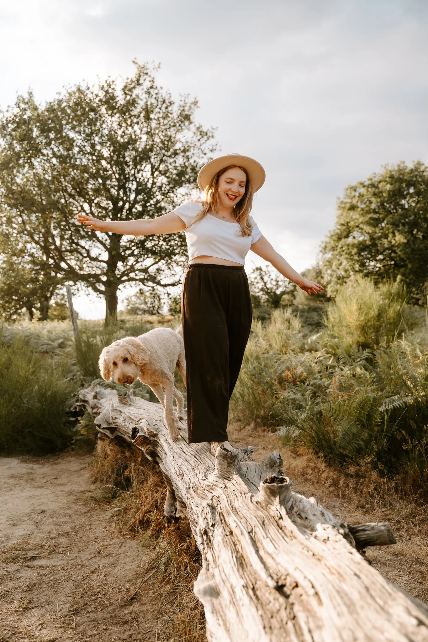 Junge Frau mit Hut, die auf einem Baumstamm in der Natur balanciert, begleitet von einem Hund, umgeben von Bäumen und Gras, bei Sonnenuntergang.