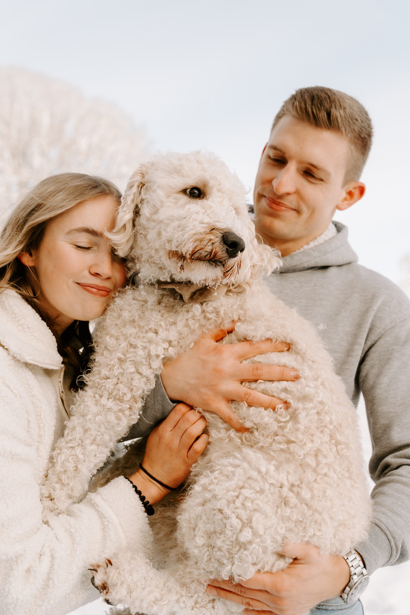 Ein junges Paar umarmt und kuschelt mit einem großen, lockigen weißen Hund im Freien an einem winterlichen Tag.