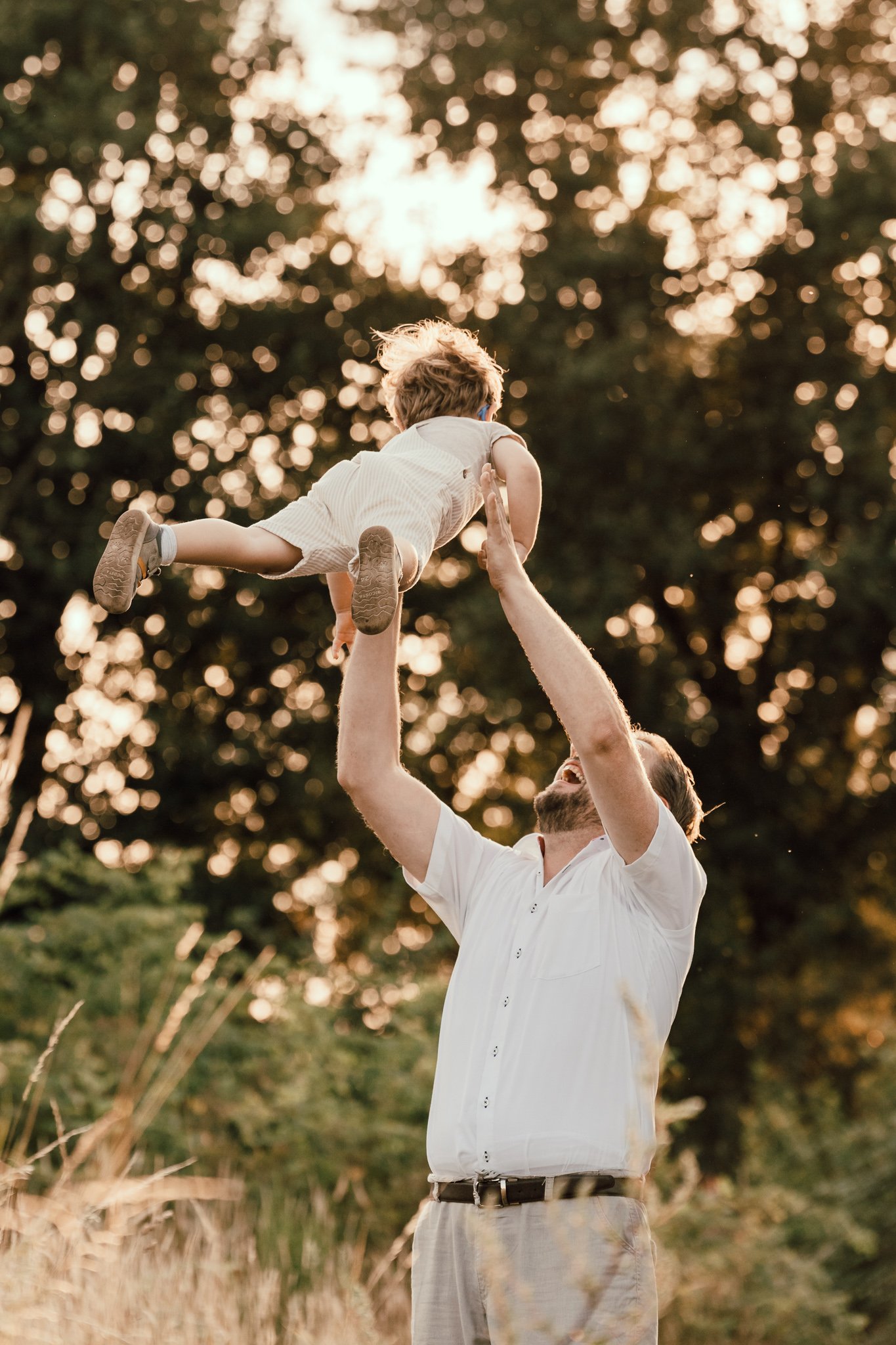 Ein Mann hebt ein Kind in einem Park bei Sonnenuntergang hoch, umgeben von Bäumen und Natur.