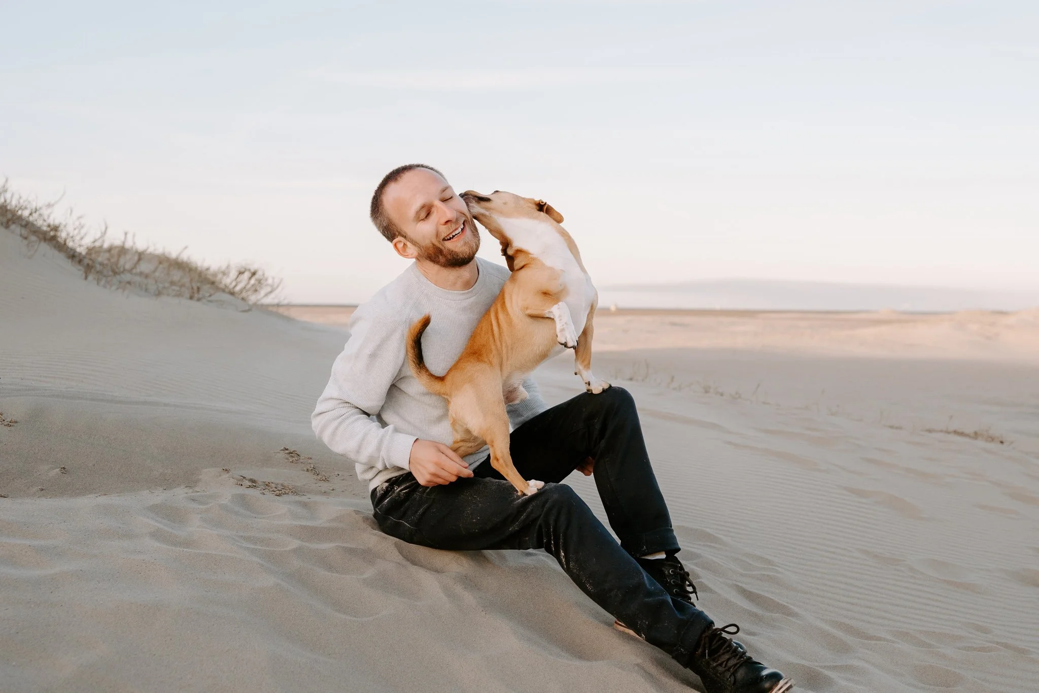 Ein lachender Mann sitzt im Sand einer Wüste und wird von einem kleinen Hund geküsst.