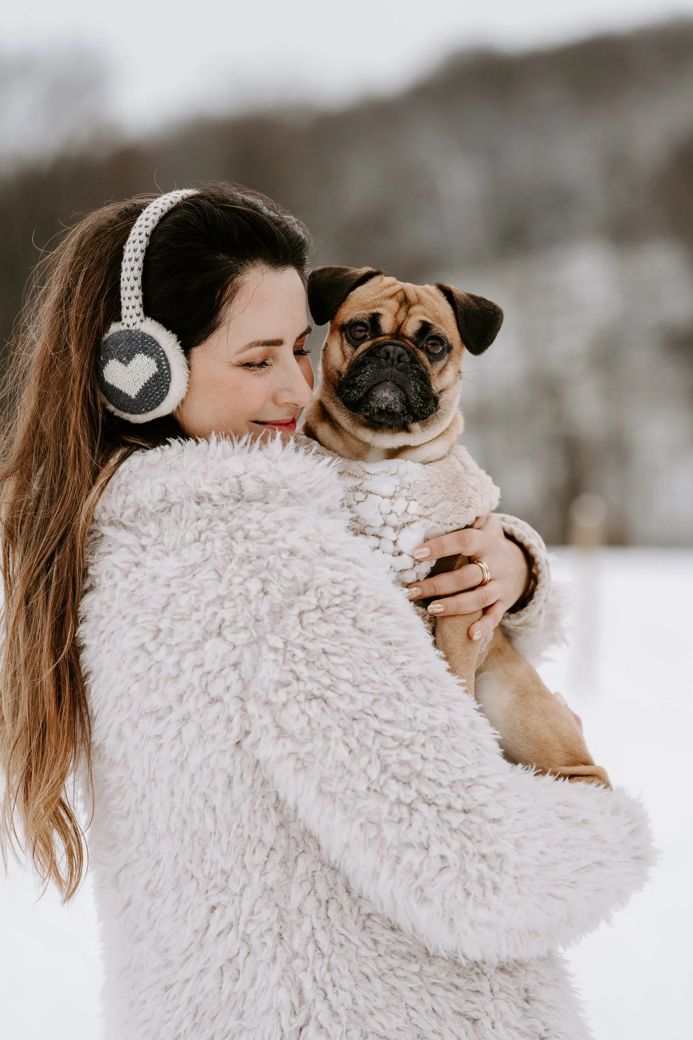 Eine lachende junge Frau mit langen braunen Haaren, trägt eine weiße, pelzige Jacke und Ohrwärmer mit Herzmuster, hält einen Bulldoggen-Hund in den Armen, während es draußen schneit.
