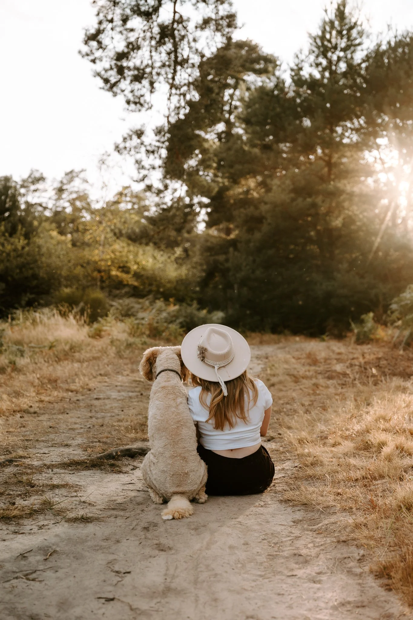 Ein Mädchen sitzt mit ihrem Hund in einer ländlichen Gegend bei Sonnenuntergang. Sie trägt einen Bohohut und sieht auf die Natur, während der Hund neben ihr sitzt.
