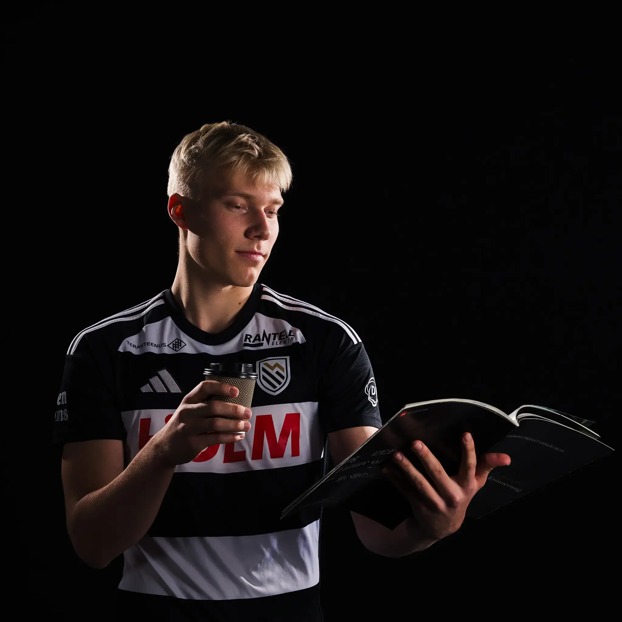 Tartu JK Welco mängija Rendo Roots poseerib stuudios portreefotoks tumedal taustal jalgpalliklubisärgis hoides käes kohvitopsi ja sirvides ajakirja