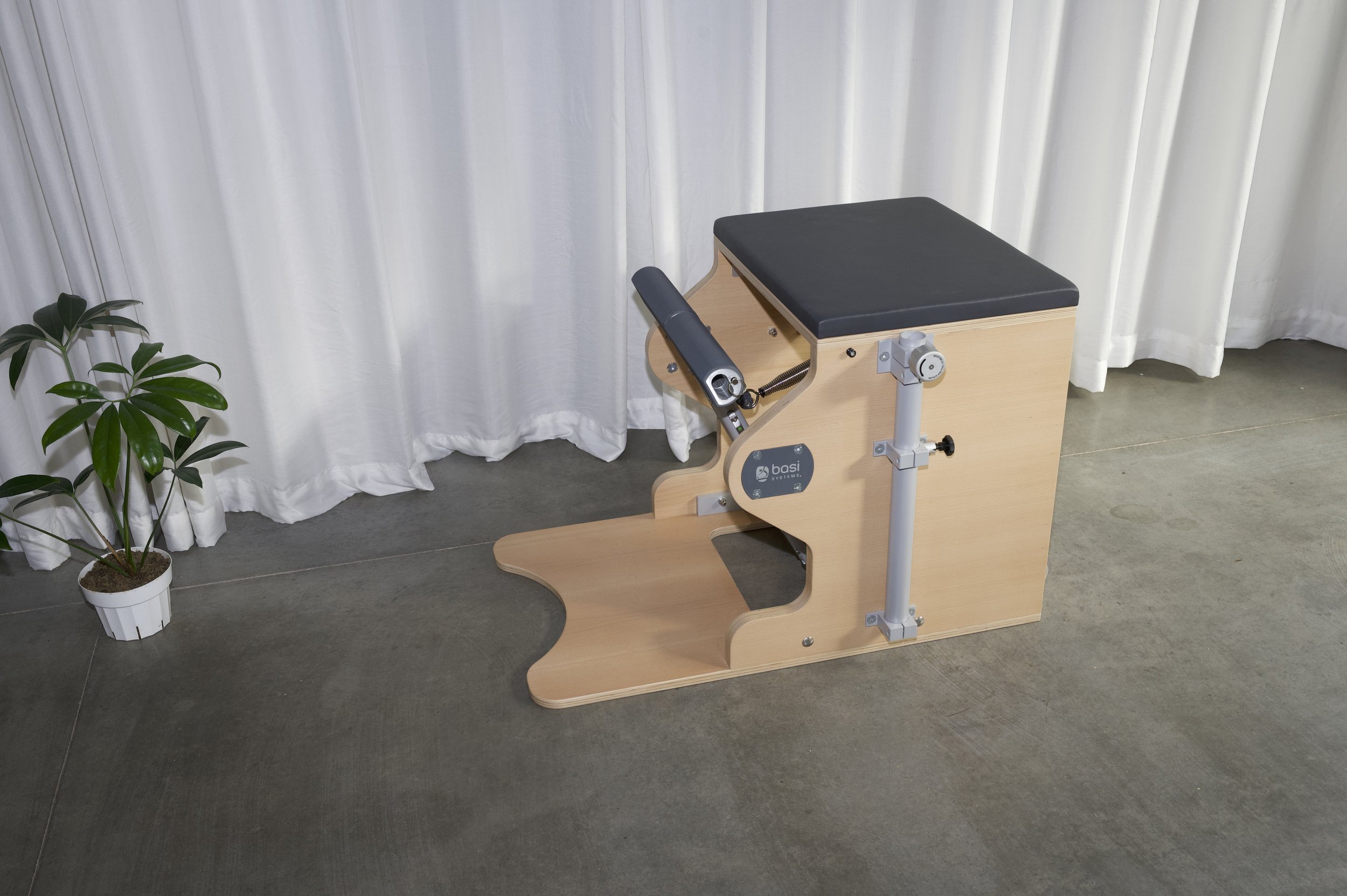 A BASI Pilates Chair with a padded seat and footrest, placed on a concrete floor with white curtains and a small potted plant nearby.