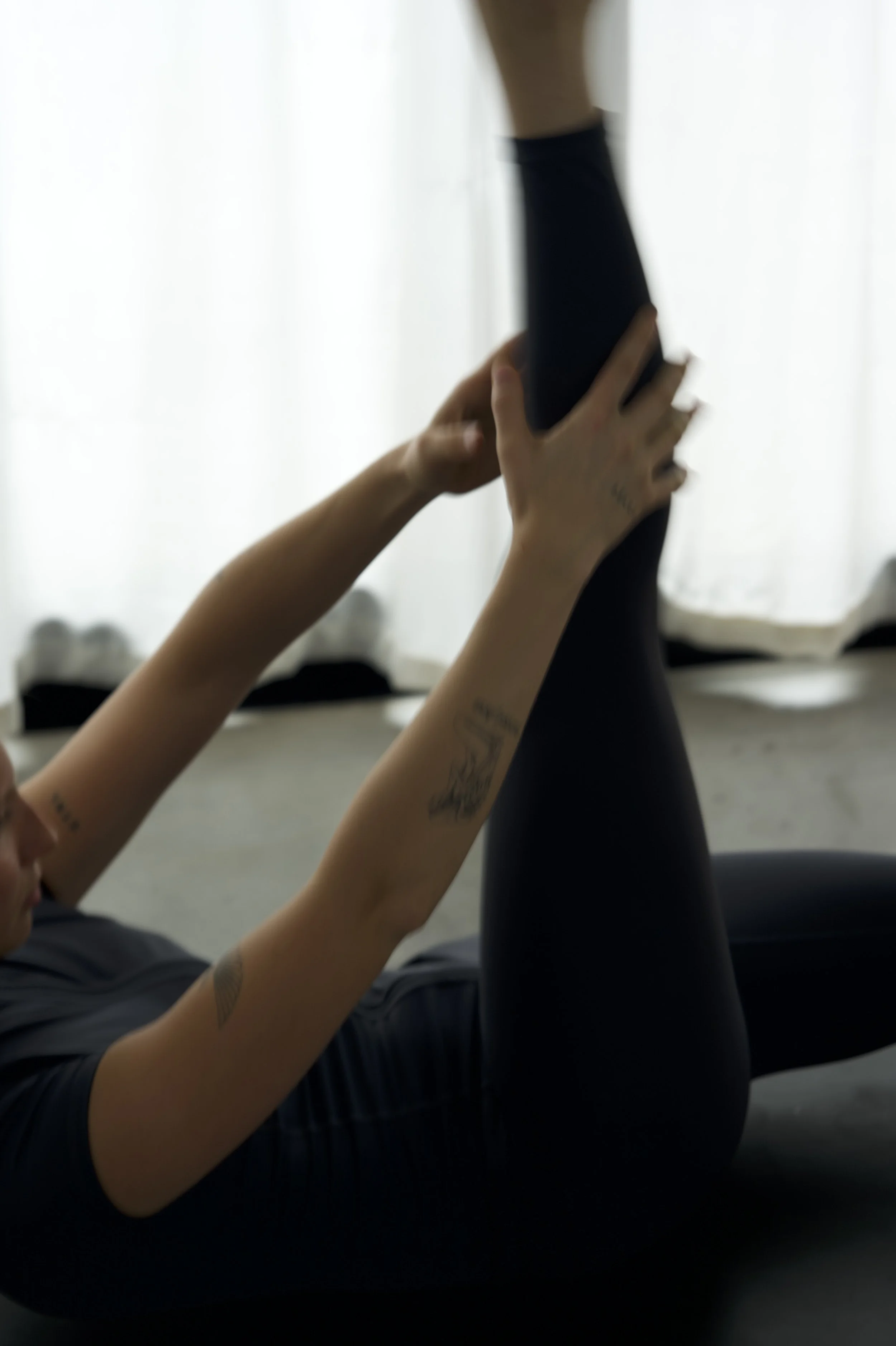 A woman lying on her back on a yoga mat, stretching one leg upward while holding it with her hands, indoors with white curtains in the background.