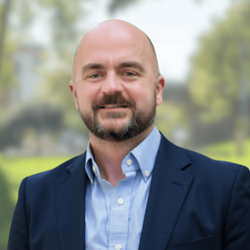 A man with a beard and bald head, wearing a dark blazer and light blue collared shirt, standing outdoors with trees and blurred background.