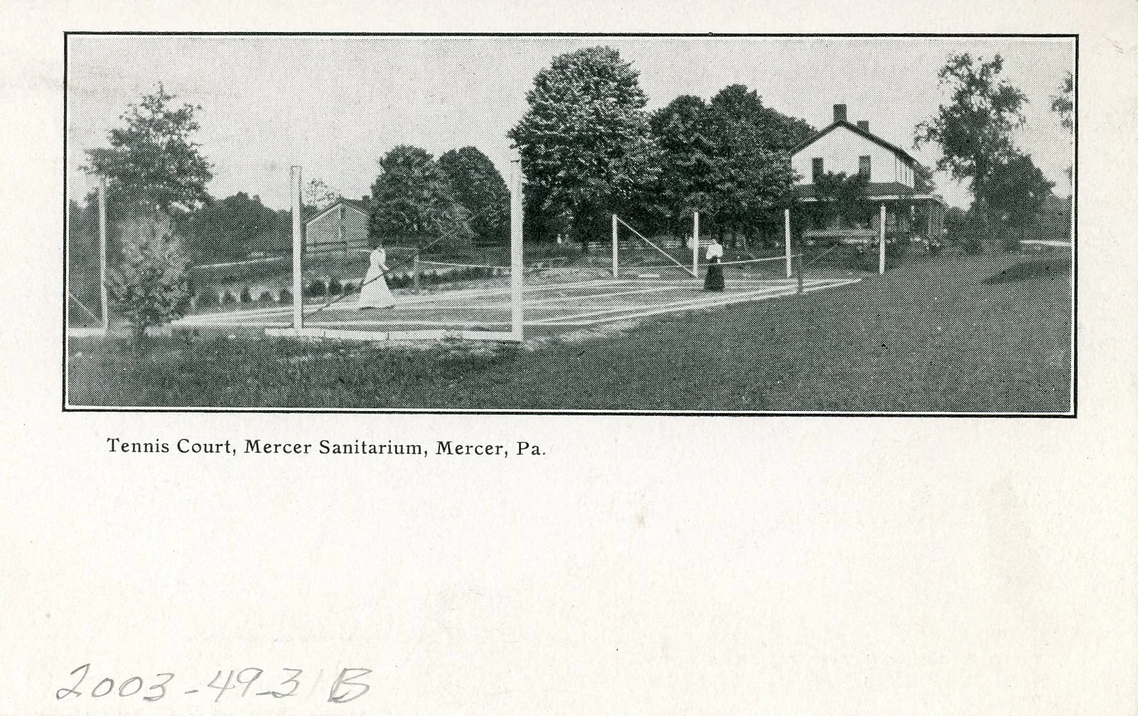 A black and white photograph of an outdoor tennis court with two women playing tennis, surrounded by trees and houses, at Mercer Sanitarium in Mercer, Pennsylvania.