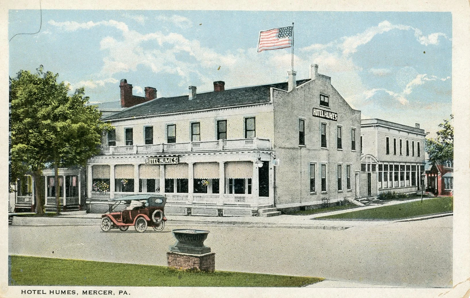 Historical photograph of the Hotel Humes in Mercer, Pennsylvania, showing a three-story white building with a sign and an American flag on top. There is an old car parked in front and a green lawn with a brick planter in the foreground. The sky is pa