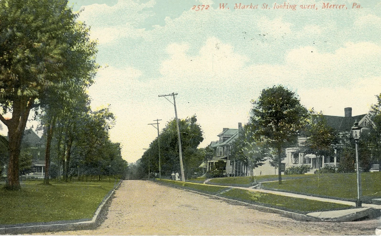 A vintage color photograph of a residential street with houses, trees, and power lines in Mercer, Pennsylvania. The street curves slightly with grassy areas and sidewalks on either side, and a few people are visible walking in the distance.