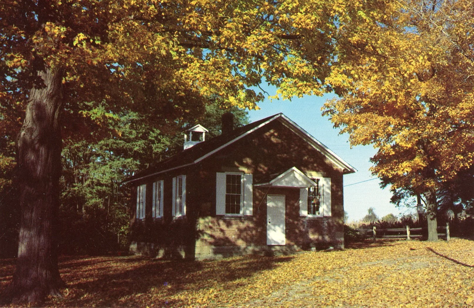 A small brick house with white window shutters, surrounded by colorful autumn trees with yellow and green leaves, on a clear day.