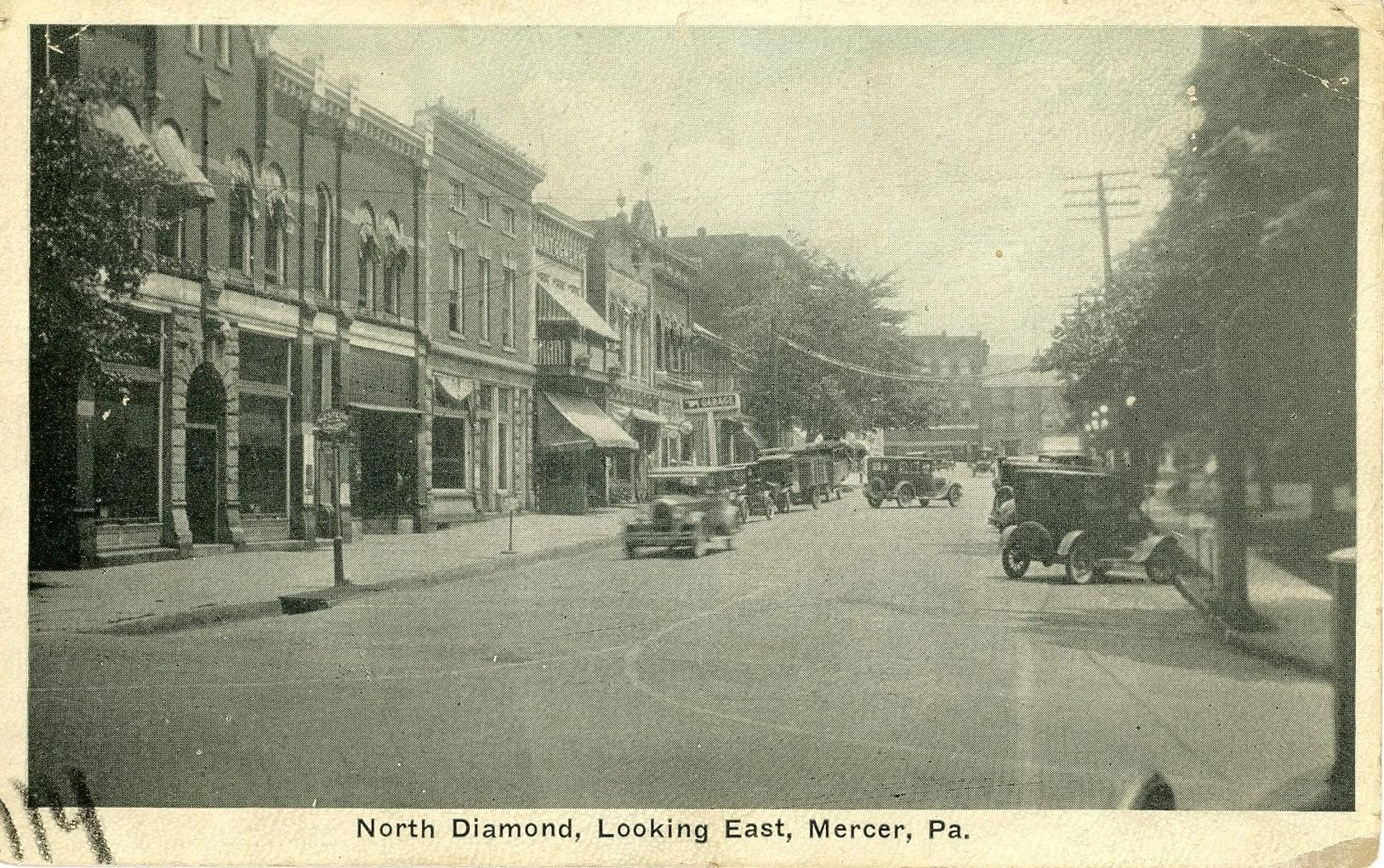 Black and white historical photograph of North Diamond Avenue, Mercer, Pennsylvania, showing a city street with old-fashioned cars parked along the sidewalk, storefronts with awnings, and trees lining the street.