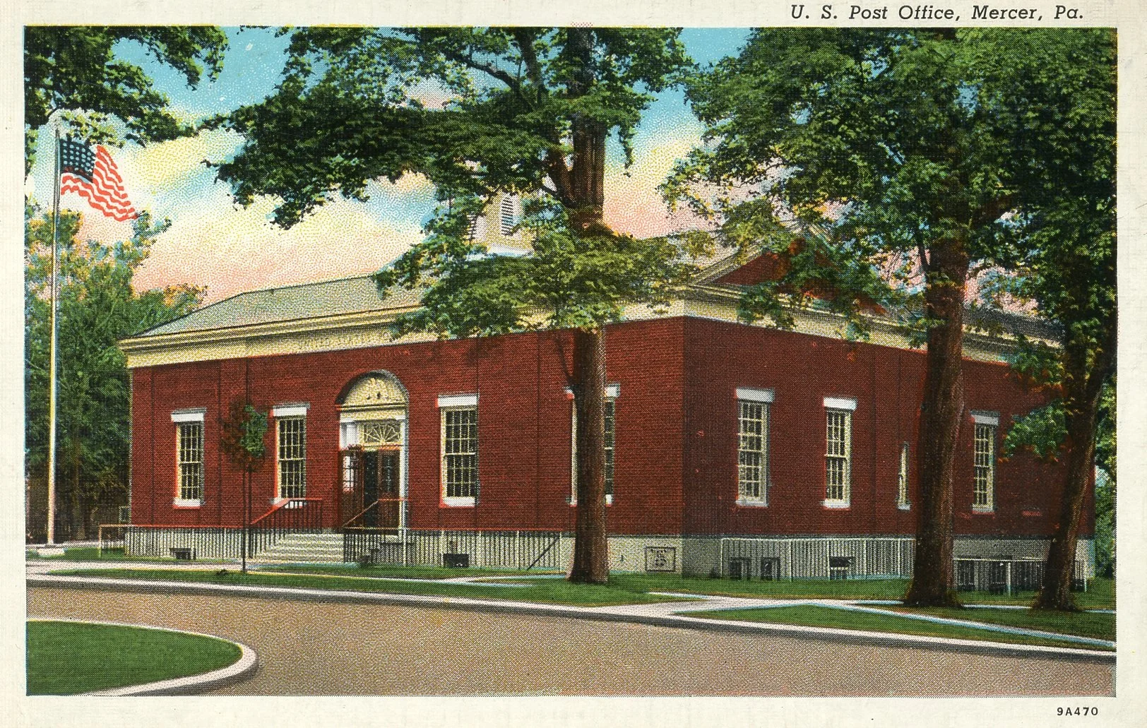 A red brick post office building with white trim, surrounded by green trees and a well-maintained lawn in Mercer, Pennsylvania. An American flag is flying on a flagpole near the building. The sky is partly cloudy with a pink and blue hue, suggesting 