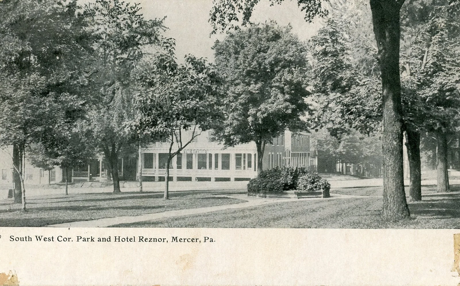 A black and white photo of a park with trees, a building in the background, and a circular flower bed with bushes in the center. The caption reads: "South West Cor. Park and Hotel Reznor, Mercer, Pa."