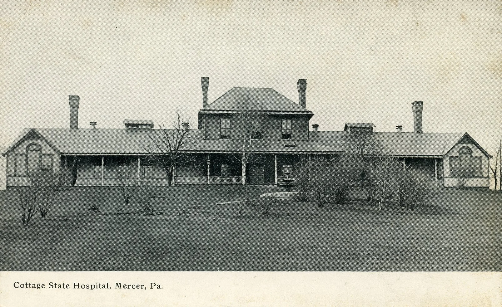 Black and white photograph of the Cottage State Hospital in Mercer, Pennsylvania, showing a large house-like building with a central section and symmetrical wings, surrounded by a lawn with small bushes and leafless trees.