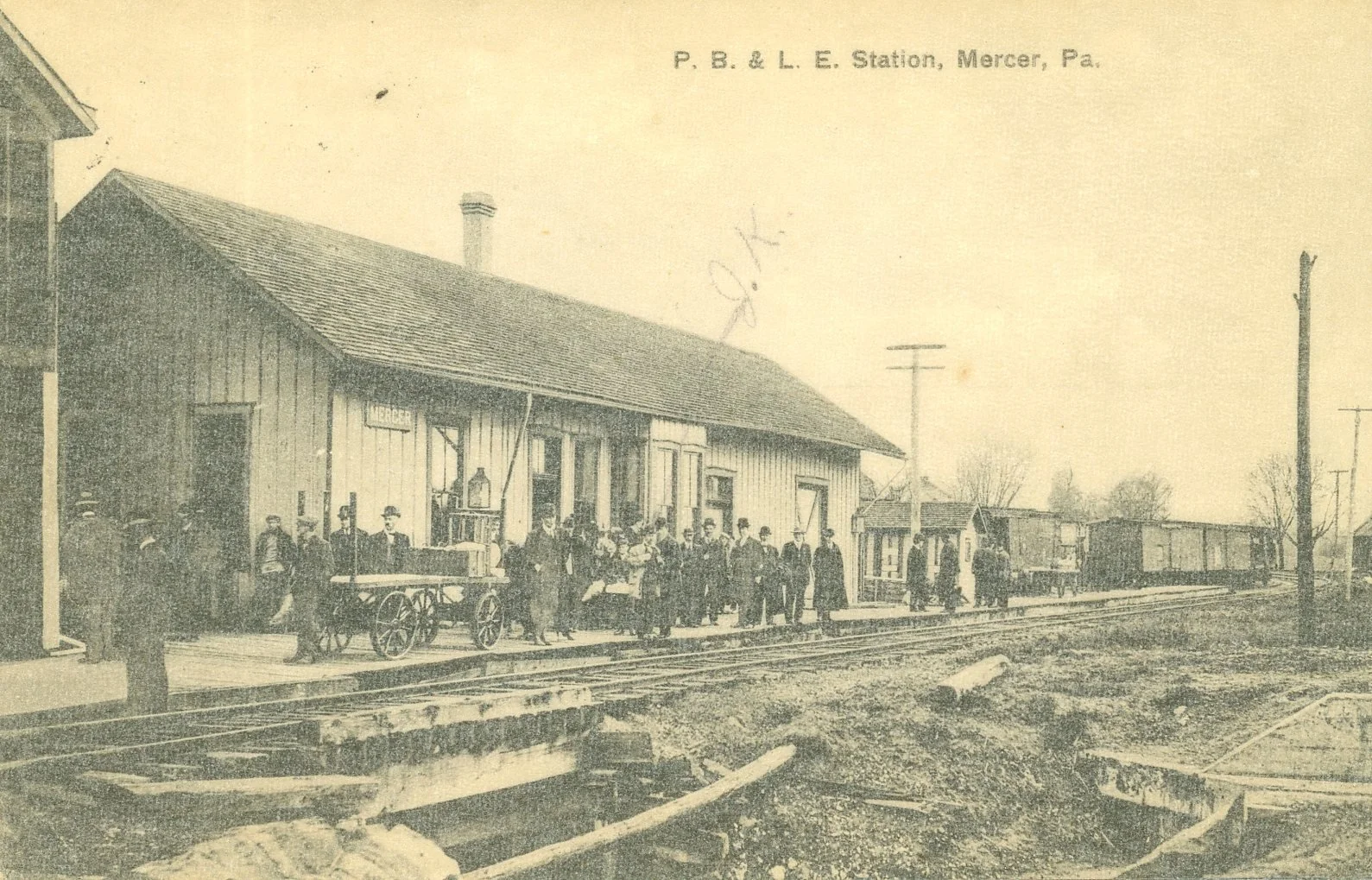 Historical black-and-white photograph of P. B. & L. E. Station in Mercer, Pennsylvania, showing a group of men and women standing on the platform in front of a wooden station building with a sign that reads "Mercer." Railroad tracks are visible in th