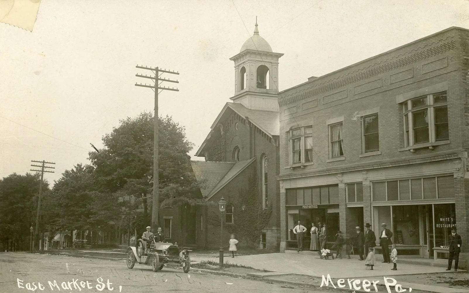 A vintage black-and-white photograph of East Market Street and Mercer Park with a church building, a post office, and several pedestrians and a car from the early 20th century.