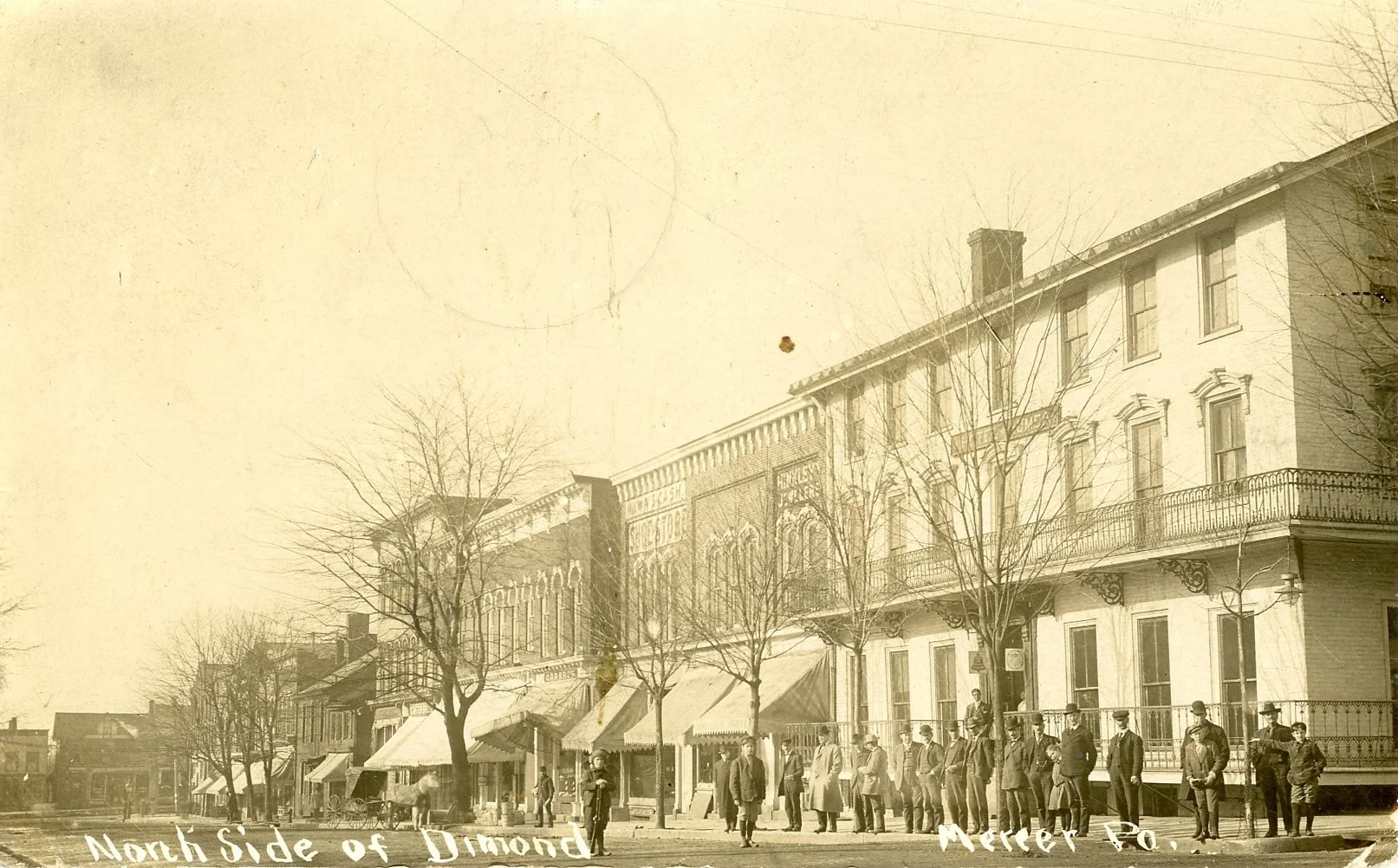Historical street scene on the north side of Diamond Street in Neffs, Pennsylvania, in the early 20th century. People dressed in period clothing stand in a line outside shops with storefront awnings, trees without leaves, and old buildings.