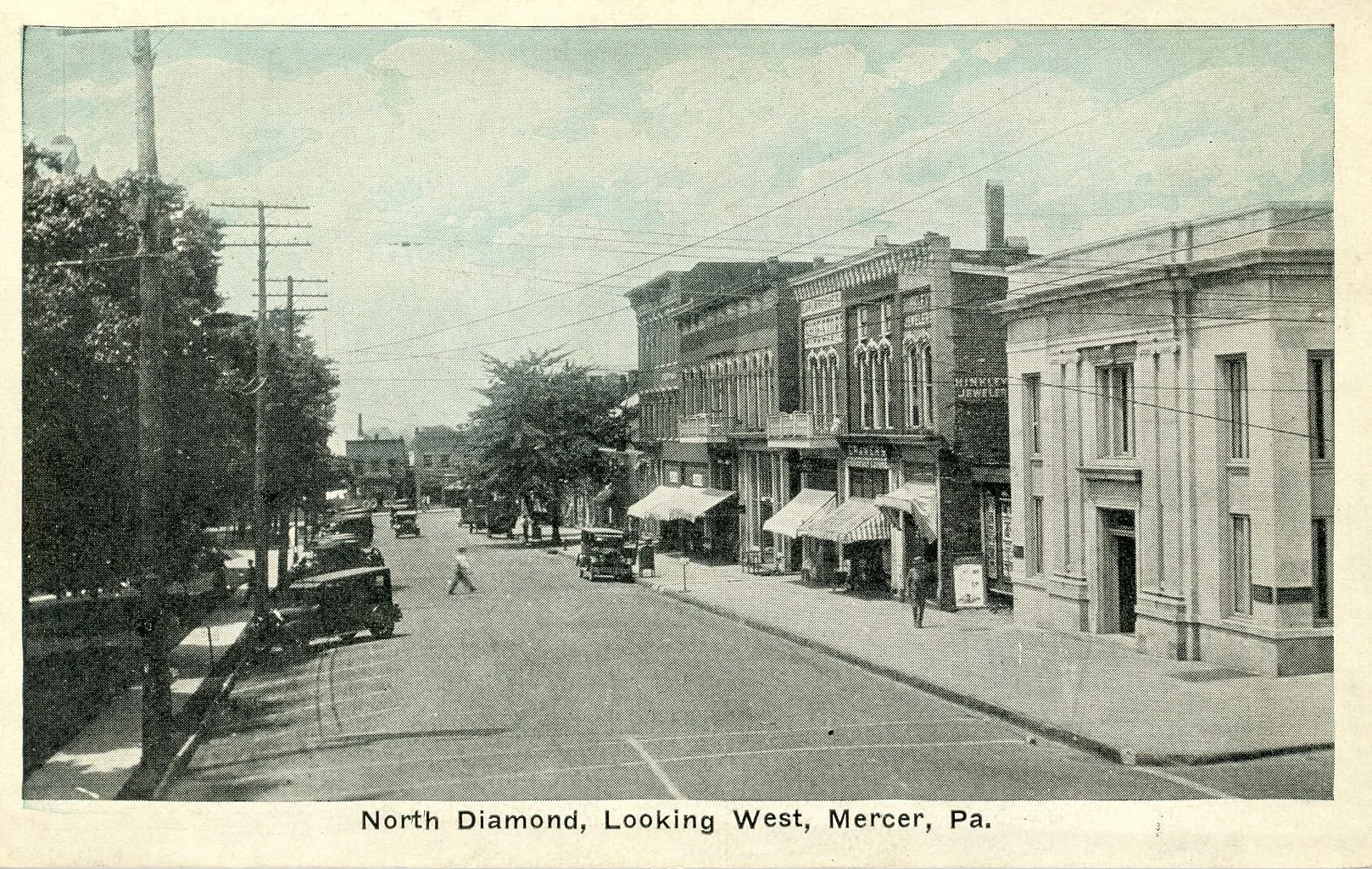 A black and white photograph of North Diamond in Mercer, Pennsylvania, looking west. The street is lined with historic brick buildings with large windows and awnings, and vintage cars parked along the street. There are a few pedestrians walking on th