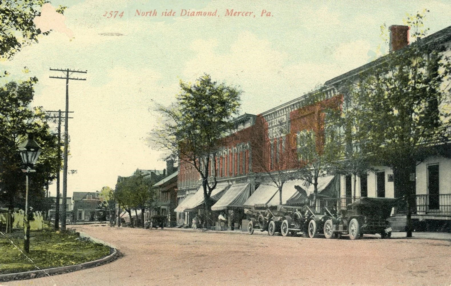 Historical street view of Mercer, Pennsylvania, featuring trees, vintage cars parked along the sidewalk, and brick buildings with storefronts under a clear sky.