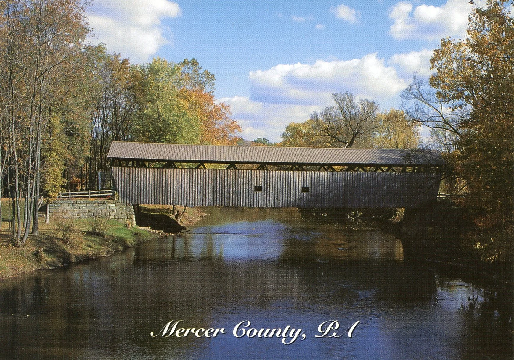 A covered bridge over a river in Mercer County, PA, with autumn foliage trees on either side and a blue sky with clouds above.