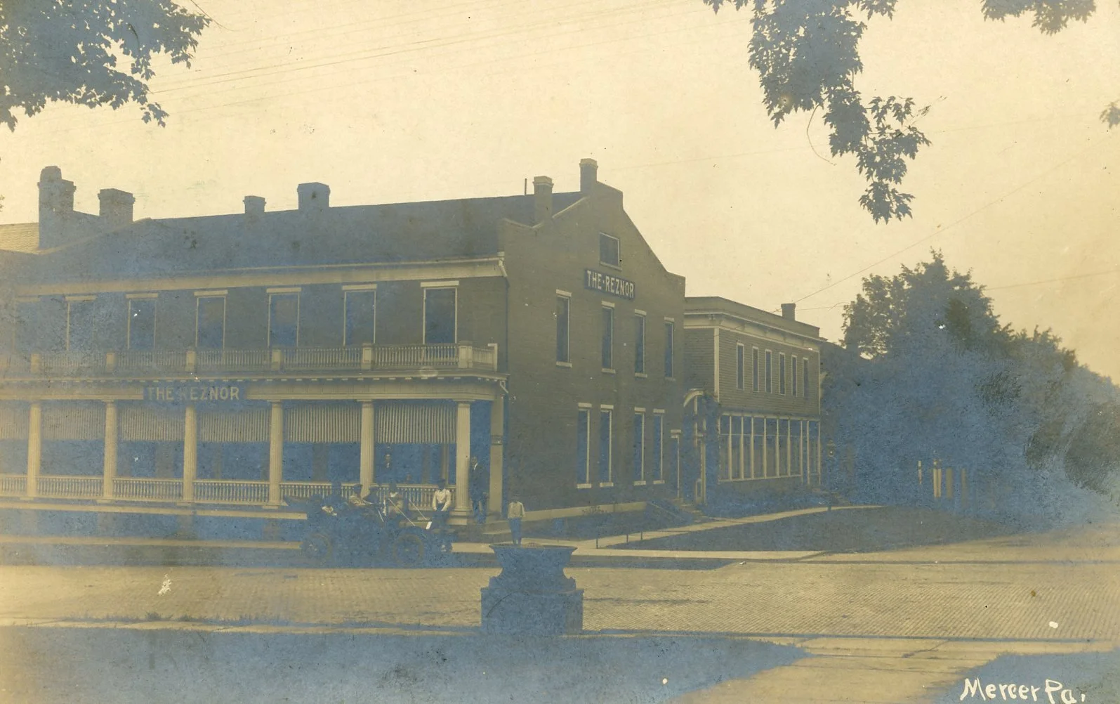 An old black-and-white photo of a three-story building labeled "The Reznor," featuring a porch with columns, a balcony, and several windows. There are a few people and vintage cars parked in front, with trees and a street in the foreground.