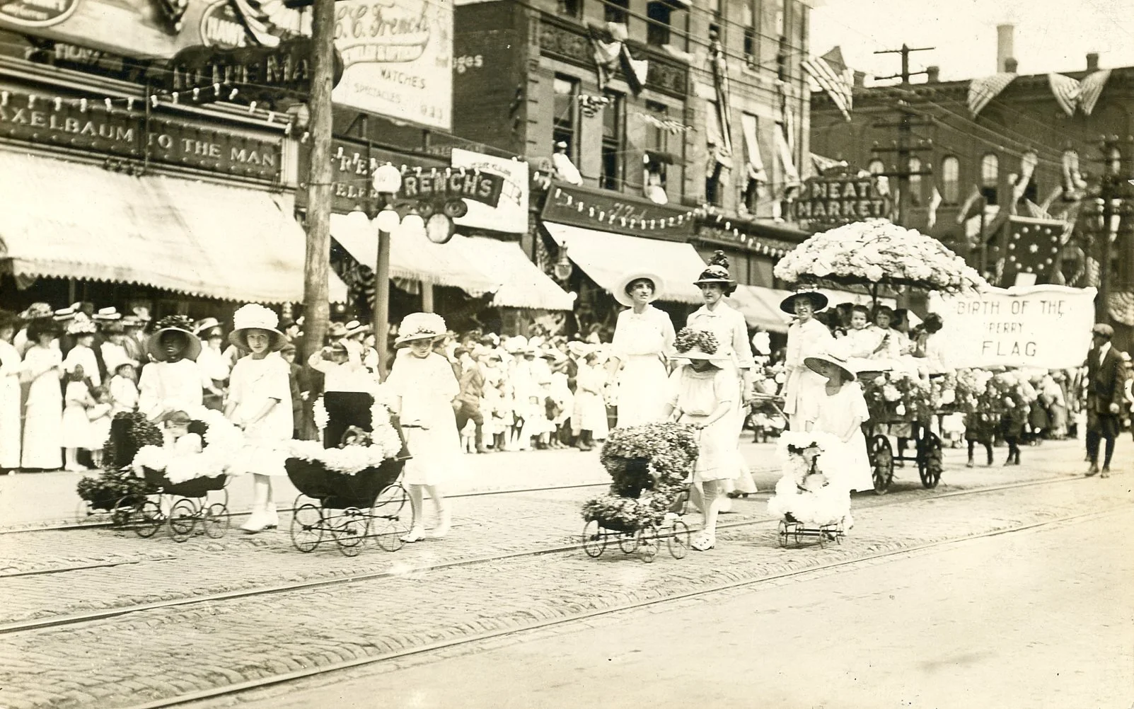 A black and white photograph of a parade on a city street, featuring young girls dressed in white and wearing large hats, walking alongside a decorated float with flowers and a sign reading 'Birth of the Perry Flag'. The float is adorned with flowers