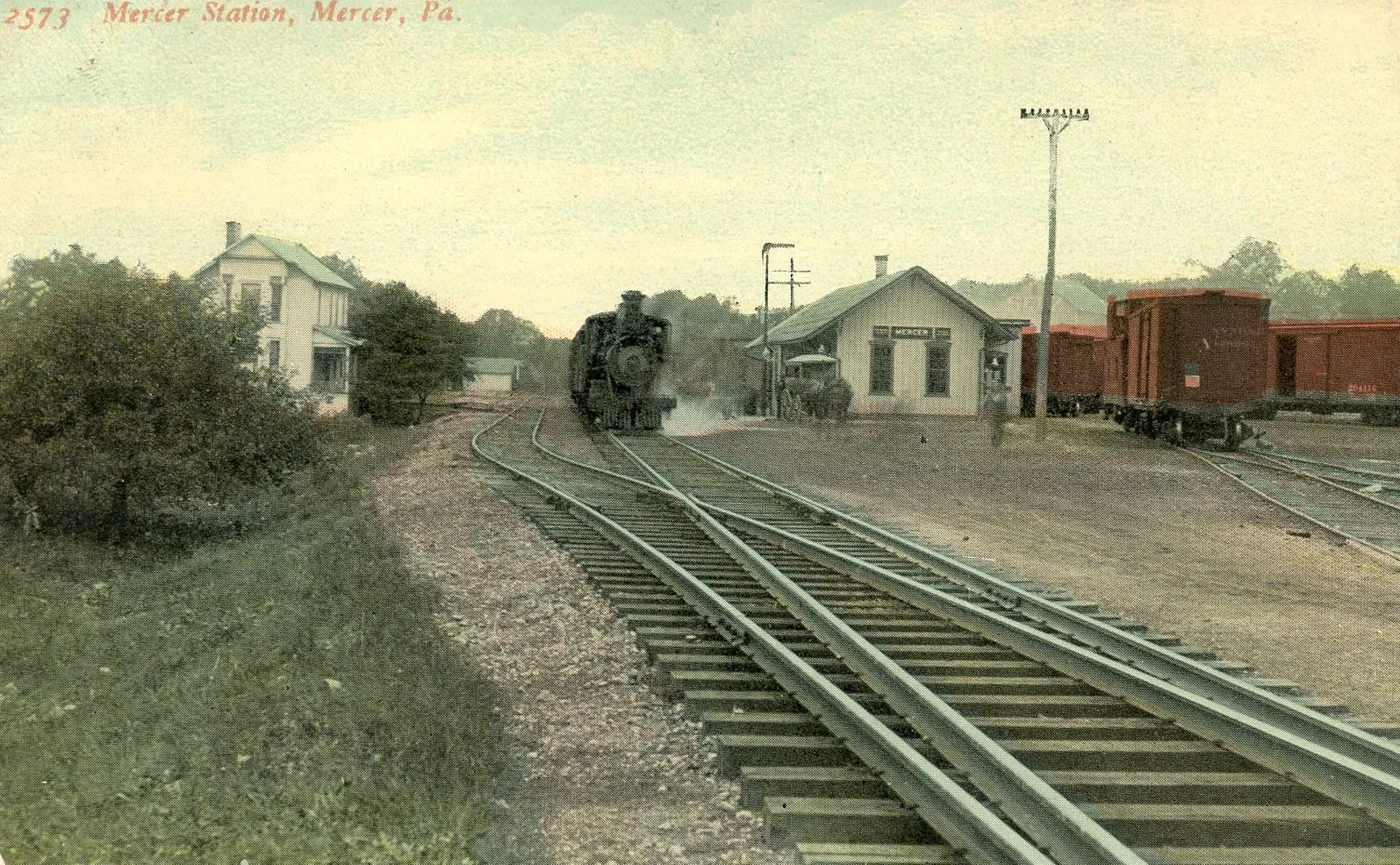 A vintage steam train arriving at Mercer Station, Mercer, Pennsylvania, near a small station building with a passenger car and freight cars on adjacent tracks.