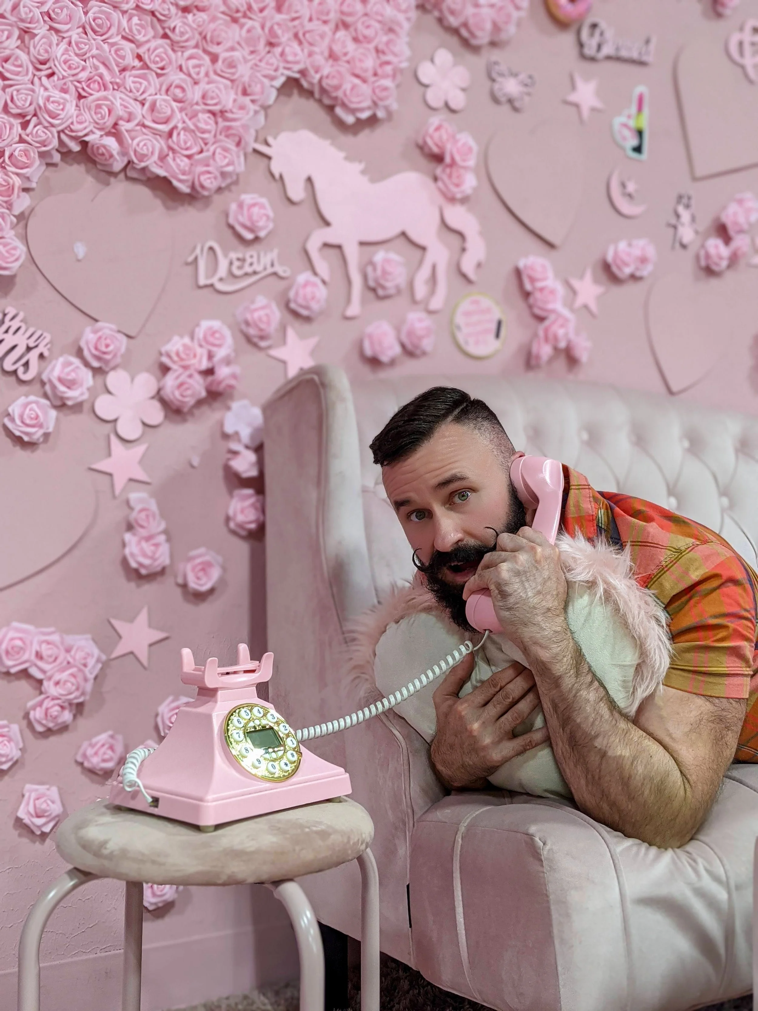 A man with a beard and vintage styled mustache lies on a pink sofa, holding a pink landline phone to his ear with one hand while a small dog rests on his chest. The background features a pink wall decorated with roses, hearts, stars, a unicorn silhouette, and the word "Dream" in pink.