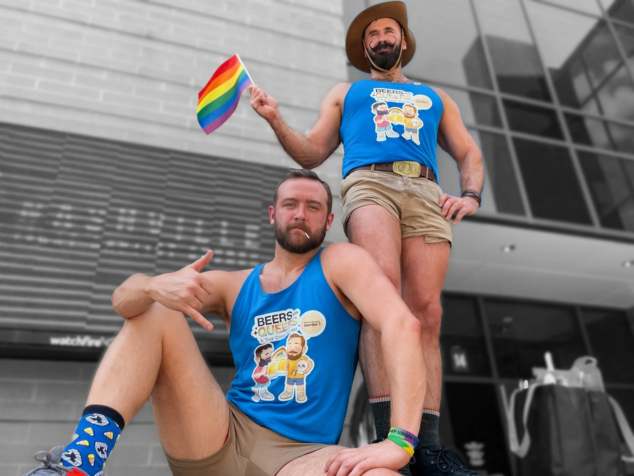 Two men wearing matching blue tank tops with LGBTQ pride theme, one standing and holding a rainbow flag, the other sitting and pointing at himself, celebrating pride outside modern building.
