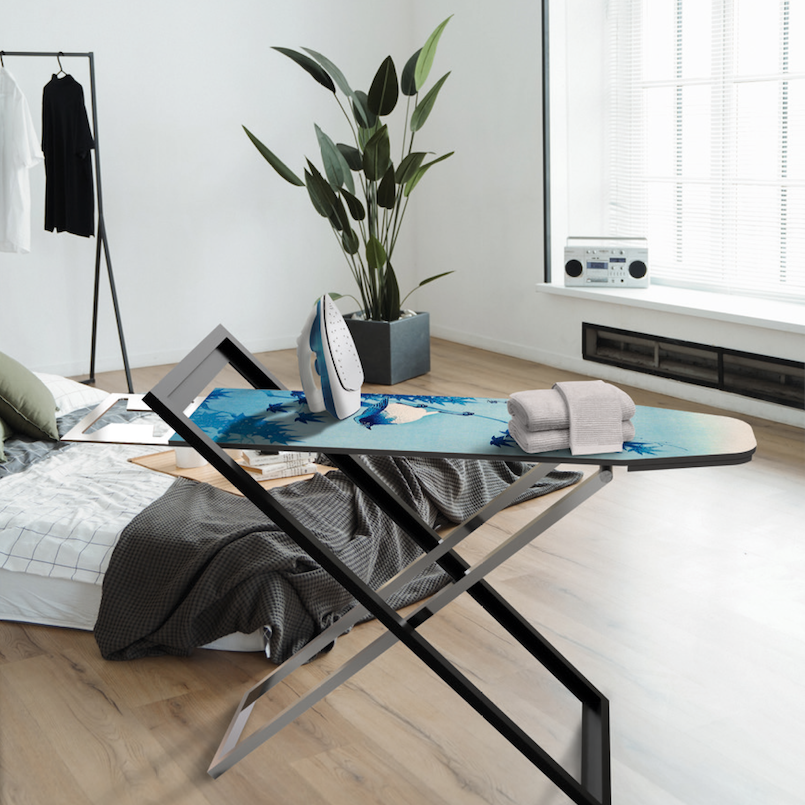 Büro- oder Wohnraum mit Bügelständer IRONY, Bett, Bügeleisen auf einem Bügeltisch, Handtücher, große Topfpflanze, Fenster mit Rollos, CD-Player.