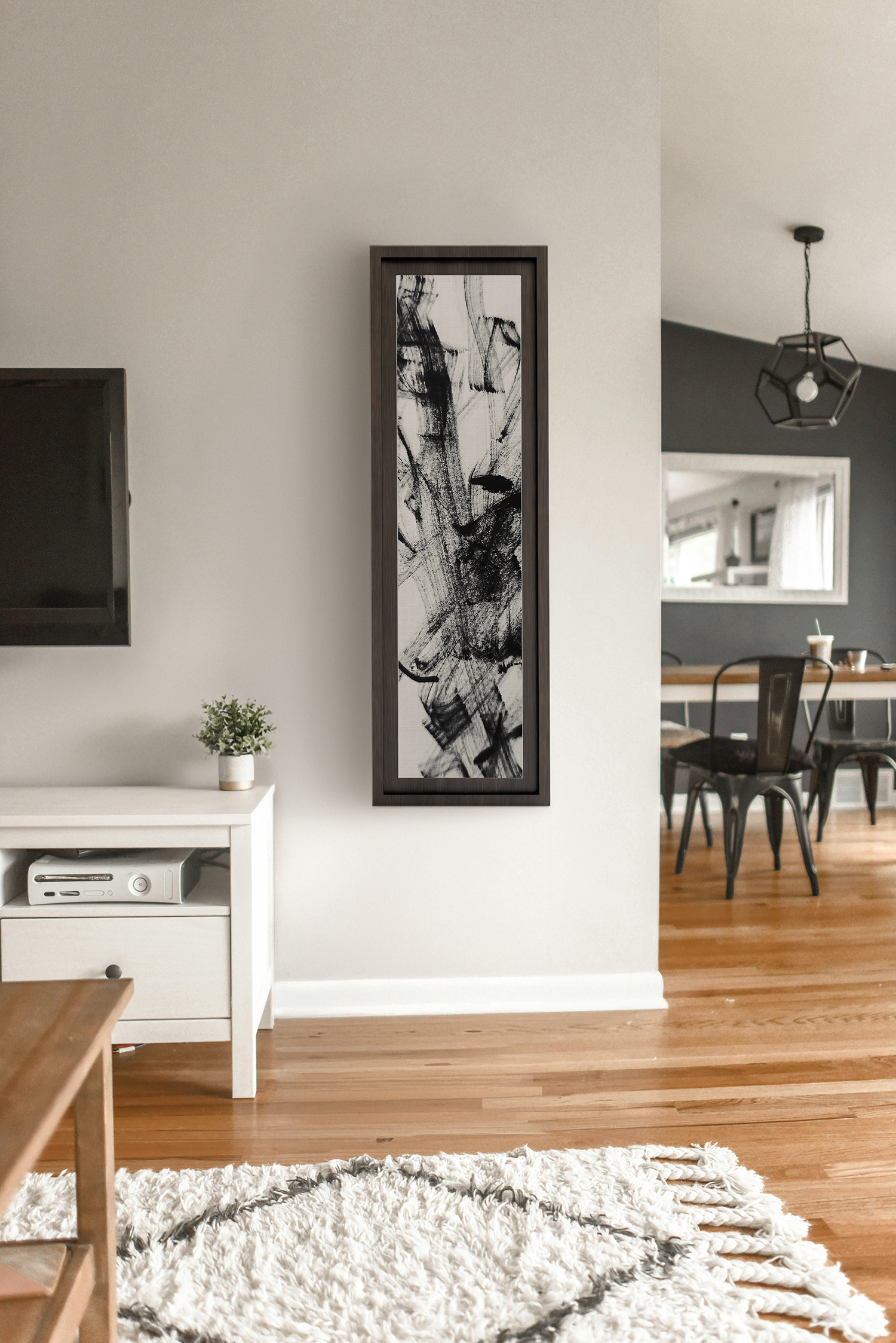 Moderner Wohnraum mit weißen Wänden, Holzboden, einem schwarzweißen Kunstwerk in einem dunklen Rahmen, einem weißen Möbelstück mit einer kleinen Pflanze und einem Fernseher, sowie einem Essbereich mit schwarzen Stühlen und einem Tisch im Hintergrund.