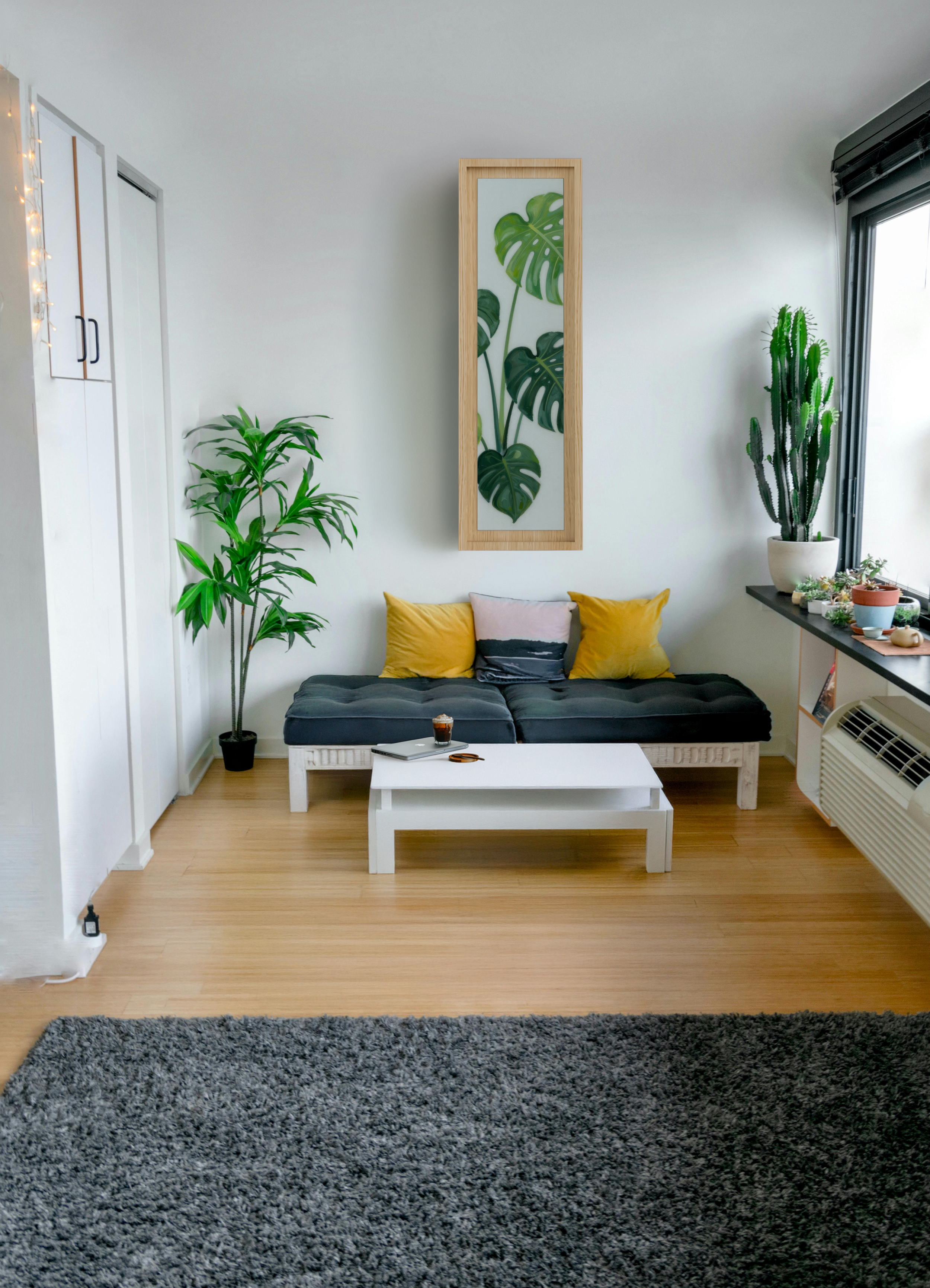 Wohnzimmer mit einer schwarzen Couch, mehreren Kissen, einem weißen Couchtisch, grünen Pflanzen und dem cleveren IRONY Bügelbrett, welches sich als dekoratives Wandbild in den hellen Raum integriert.