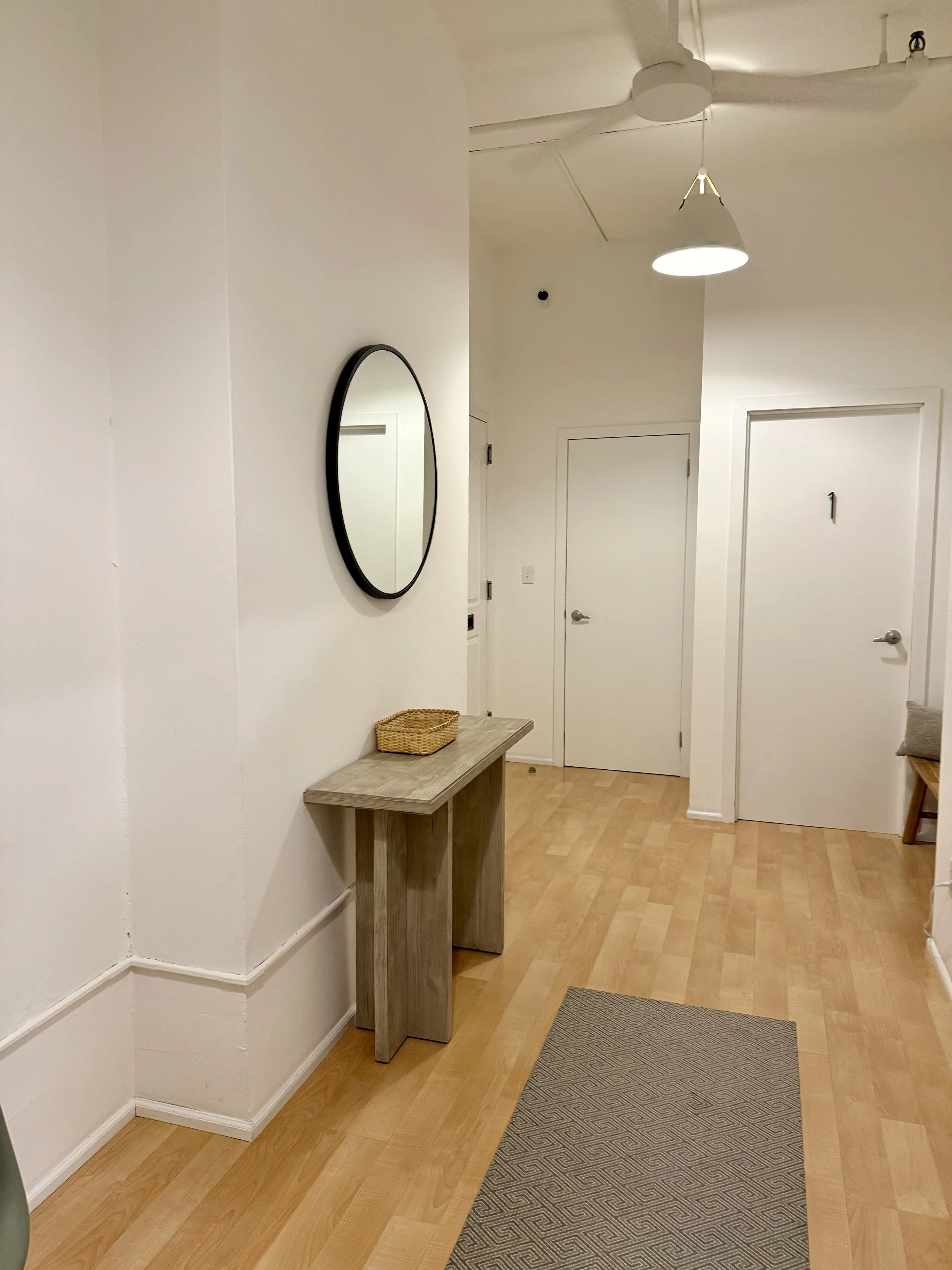 Shruthi a South Asian anxiety therapist NYC waiting area which includes a interior hallway with light-colored walls, a small wooden table with a woven basket on top, a round mirror on the wall, white doors, a ceiling fan, and a hanging light fixture.