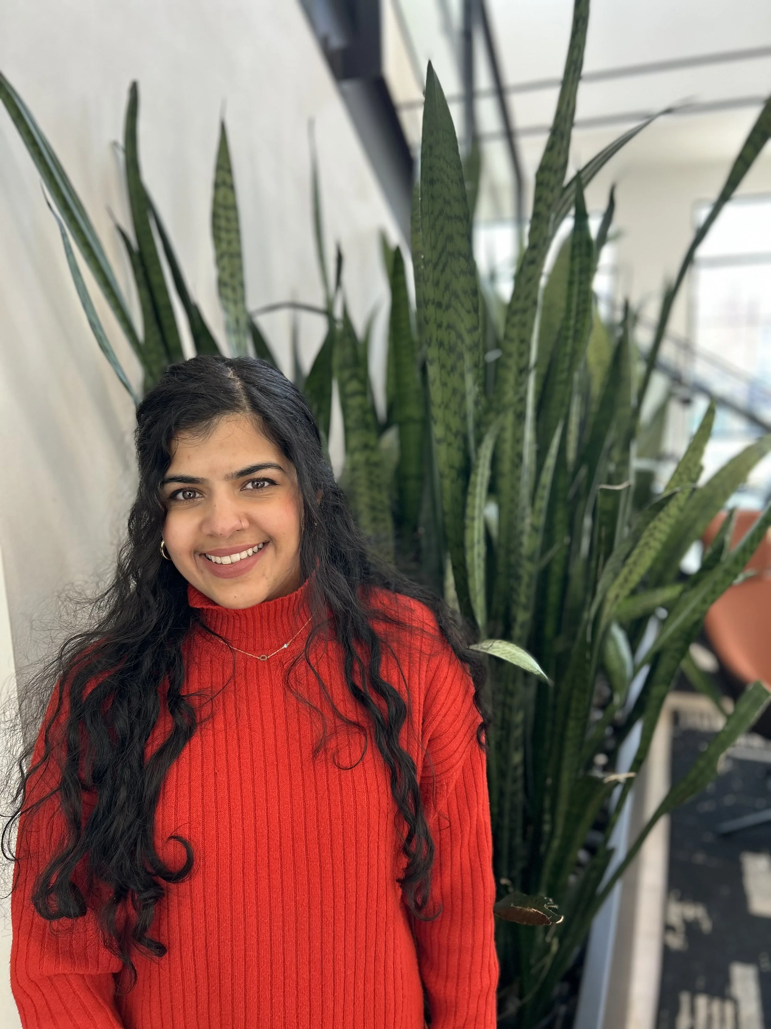 A woman with long dark curly hair and a nose piercing, smiling and wearing a red sweater, standing in front of a large green snake plant with tall, pointed leaves, inside a well-lit room with windows.