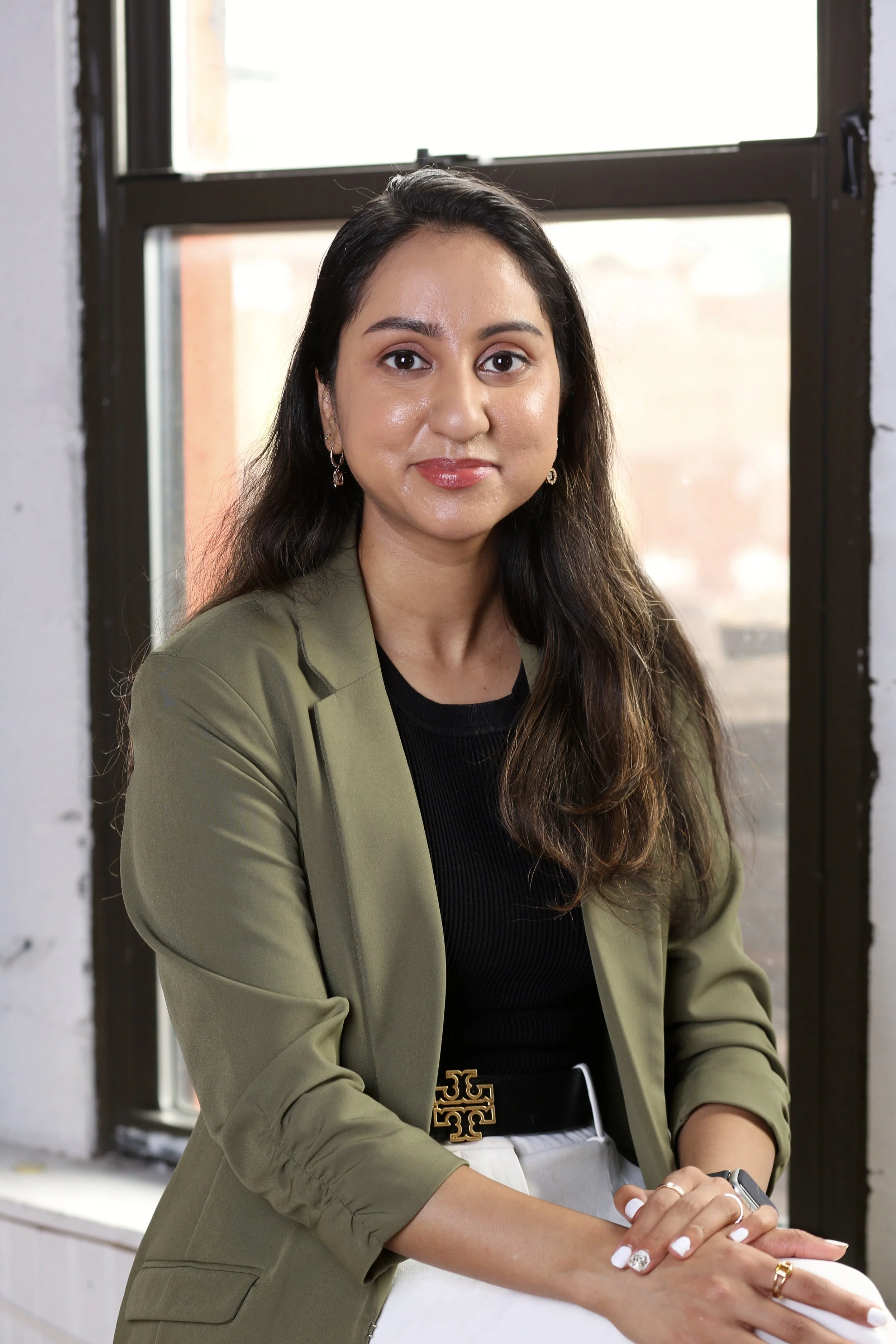 South Asian Licensed Mental Health Therapist and Founder of Bold Brown Therapy with long dark hair, wearing a light green blazer and black top, sitting by a window with a brick wall visible outside.