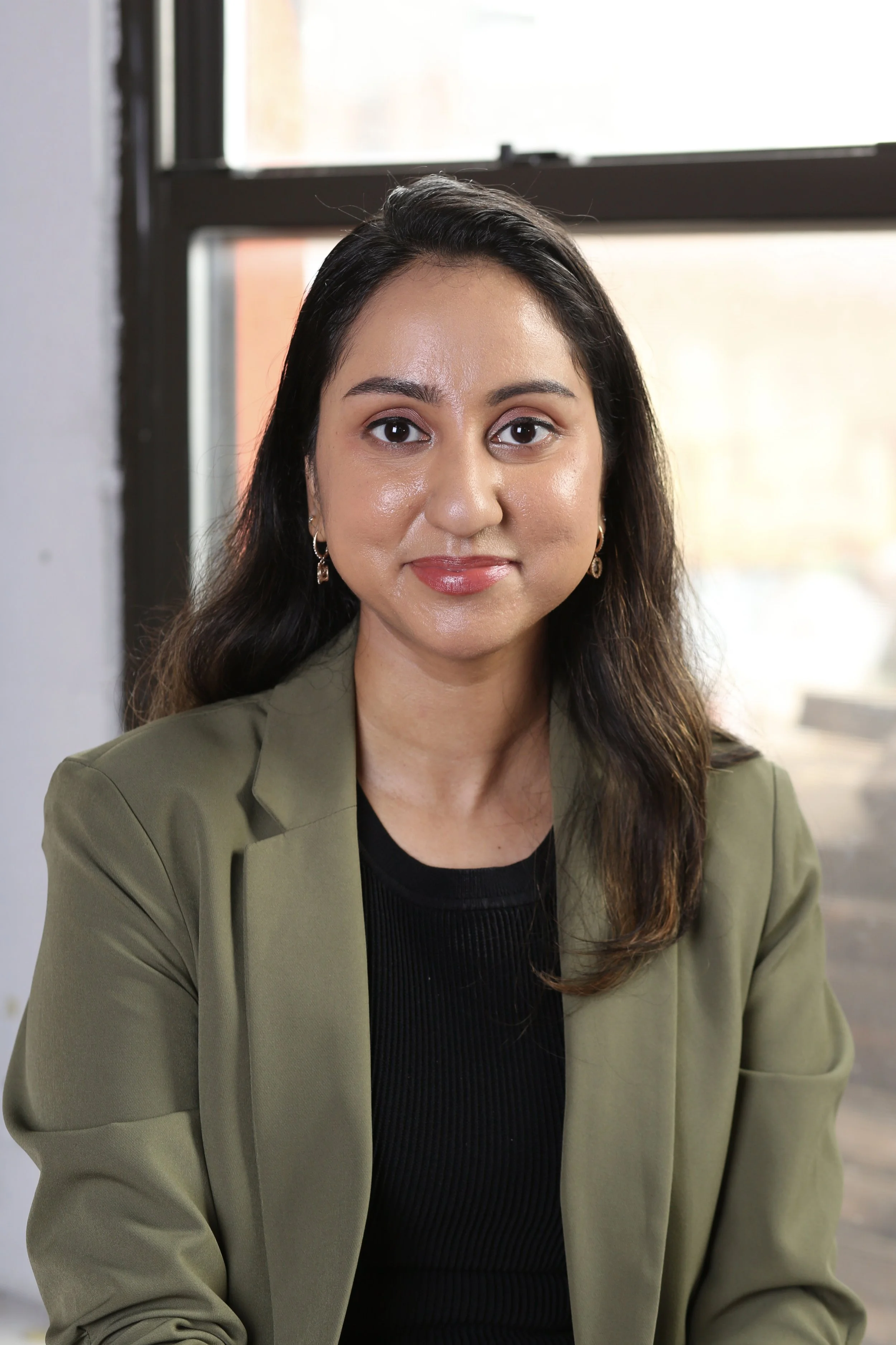 South Asian Licensed Mental Health Therapist and Founder of Bold Brown Therapy with long dark brown hair, wearing a green blazer and black top, sitting indoors near a window with a neutral background.