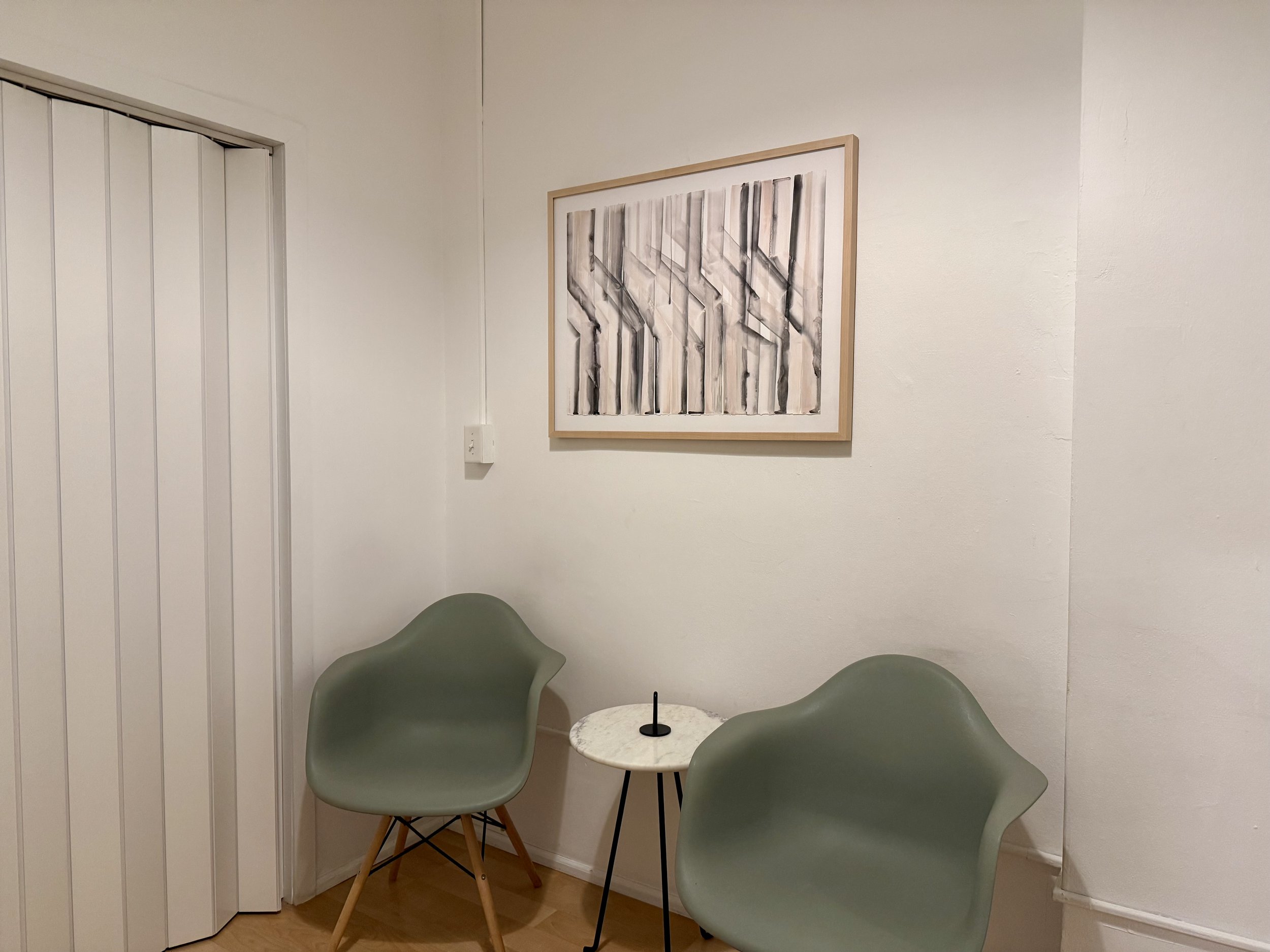 Shruthi a South Asian anxiety therapist's NYC small waiting/seating area with two green modern chairs, a white round table with a black straw, and a beige-framed abstract art piece on a white wall.