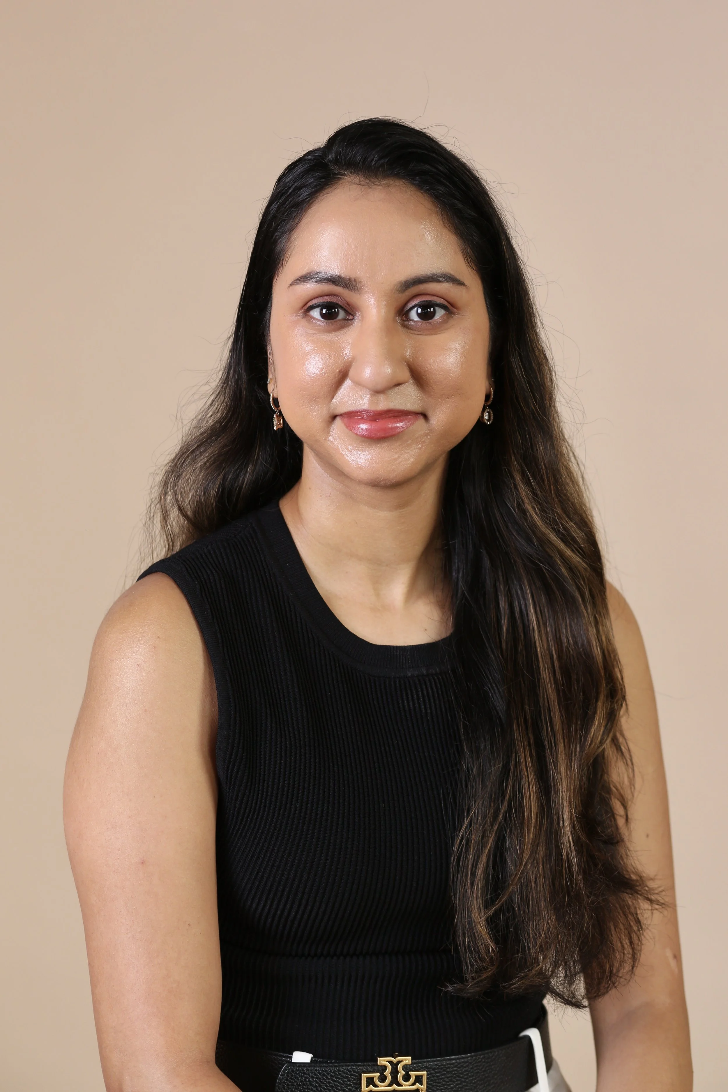 South Asian Licensed Mental Health Therapist and Founder of Bold Brown Therapy with long dark hair, wearing a black sleeveless top and earrings, smiling at the camera against a beige background.