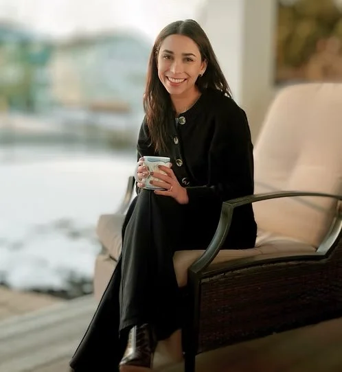 A woman sitting on a chair, holding a mug, smiling at the camera.