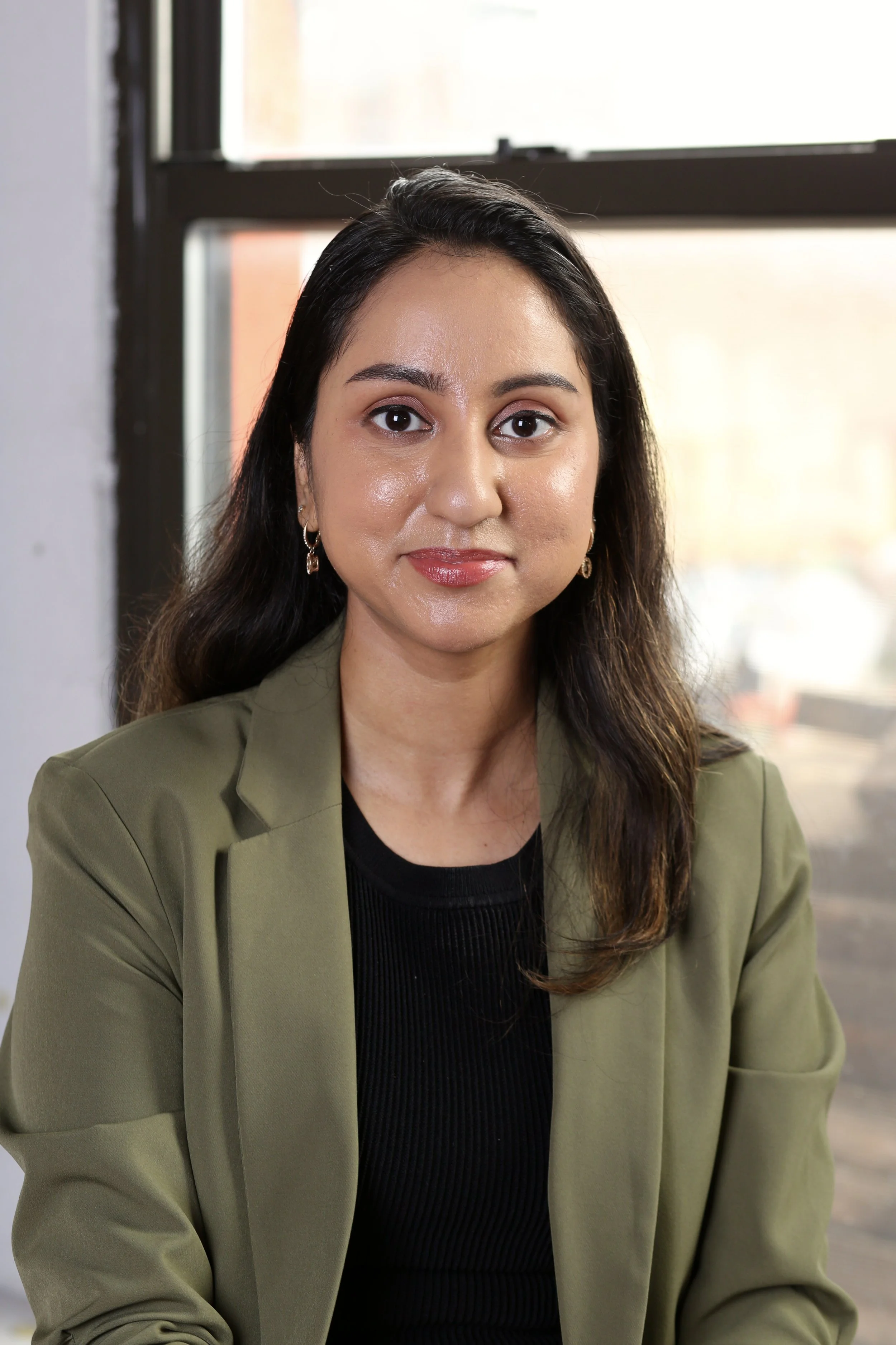 A south asian anxiety people pleasing therapist in New York City with long dark hair and wearing earrings looks directly at the camera. She is dressed in a black top and olive green blazer. The background shows a window with daylight coming through.